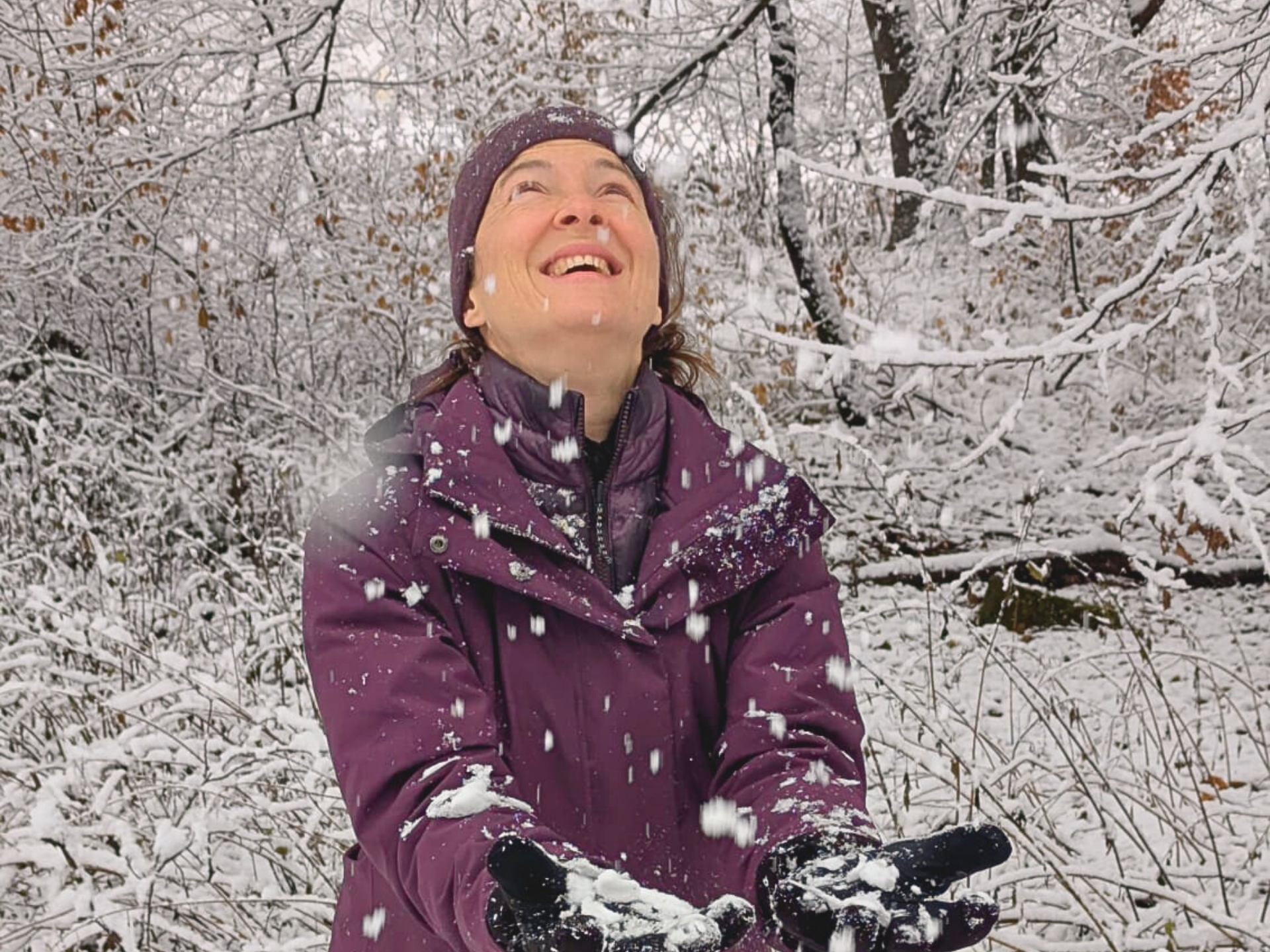 Eine Person in einem violetten Mantel lächelt und blickt nach oben, während im verschneiten Wald Schnee fällt.
