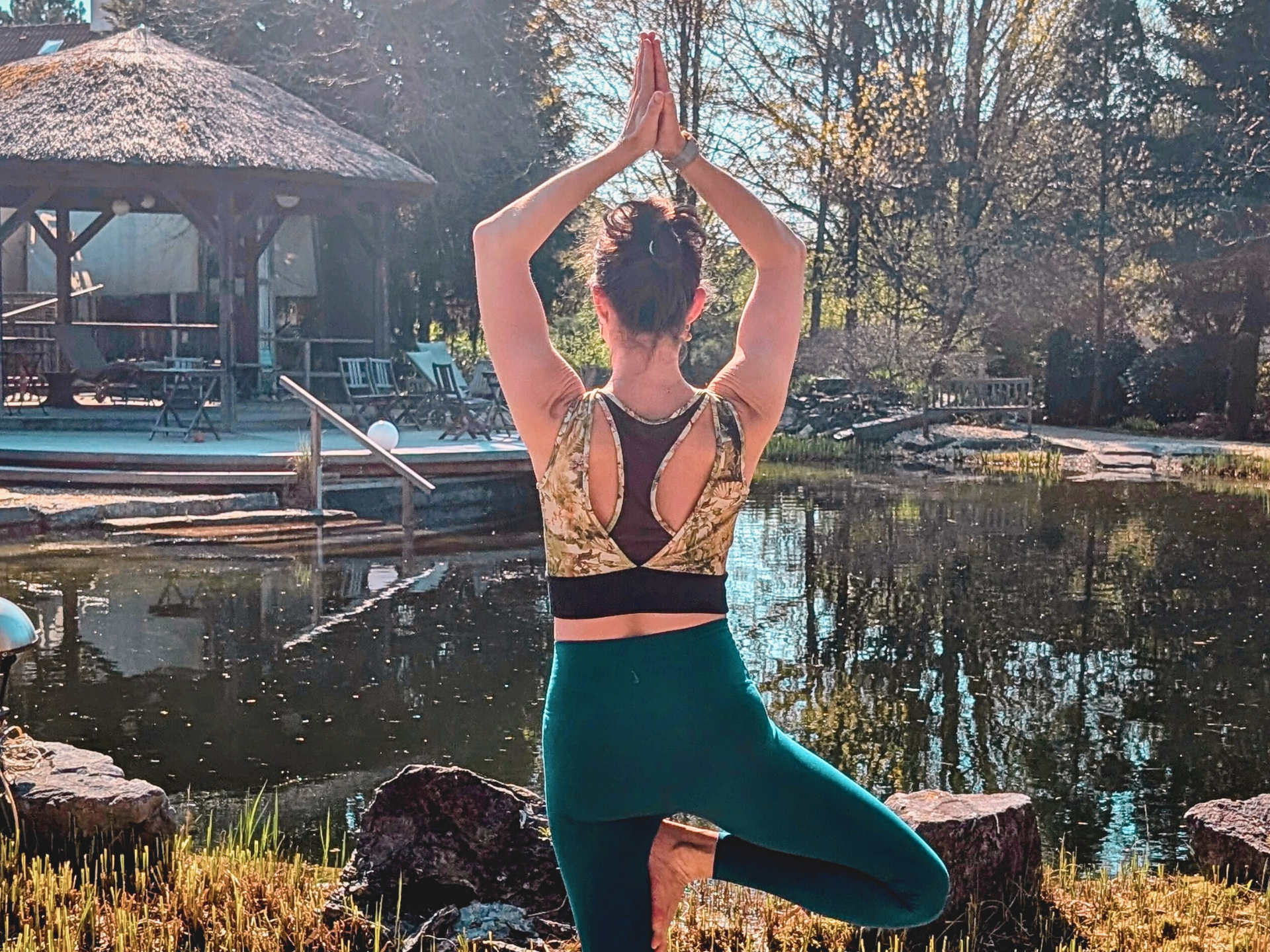 Person in der Yoga-Baumpose an einem Pool, mit über dem Kopf erhobenen Armen und einem angehobenen Bein, in einer sonnigen Umgebung im Freien