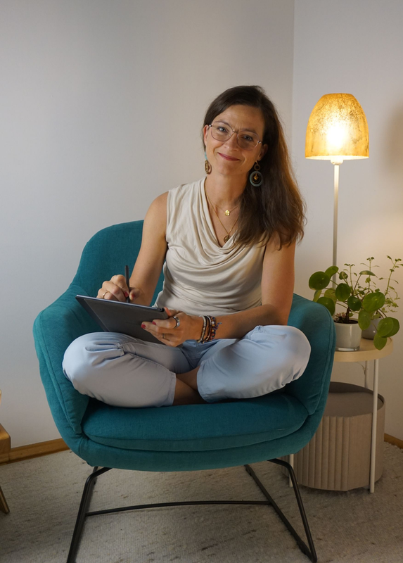 Eine lächelnde Frau sitzt mit einem Notizbuch in einem hellen Zimmer neben einer Topfpflanze.