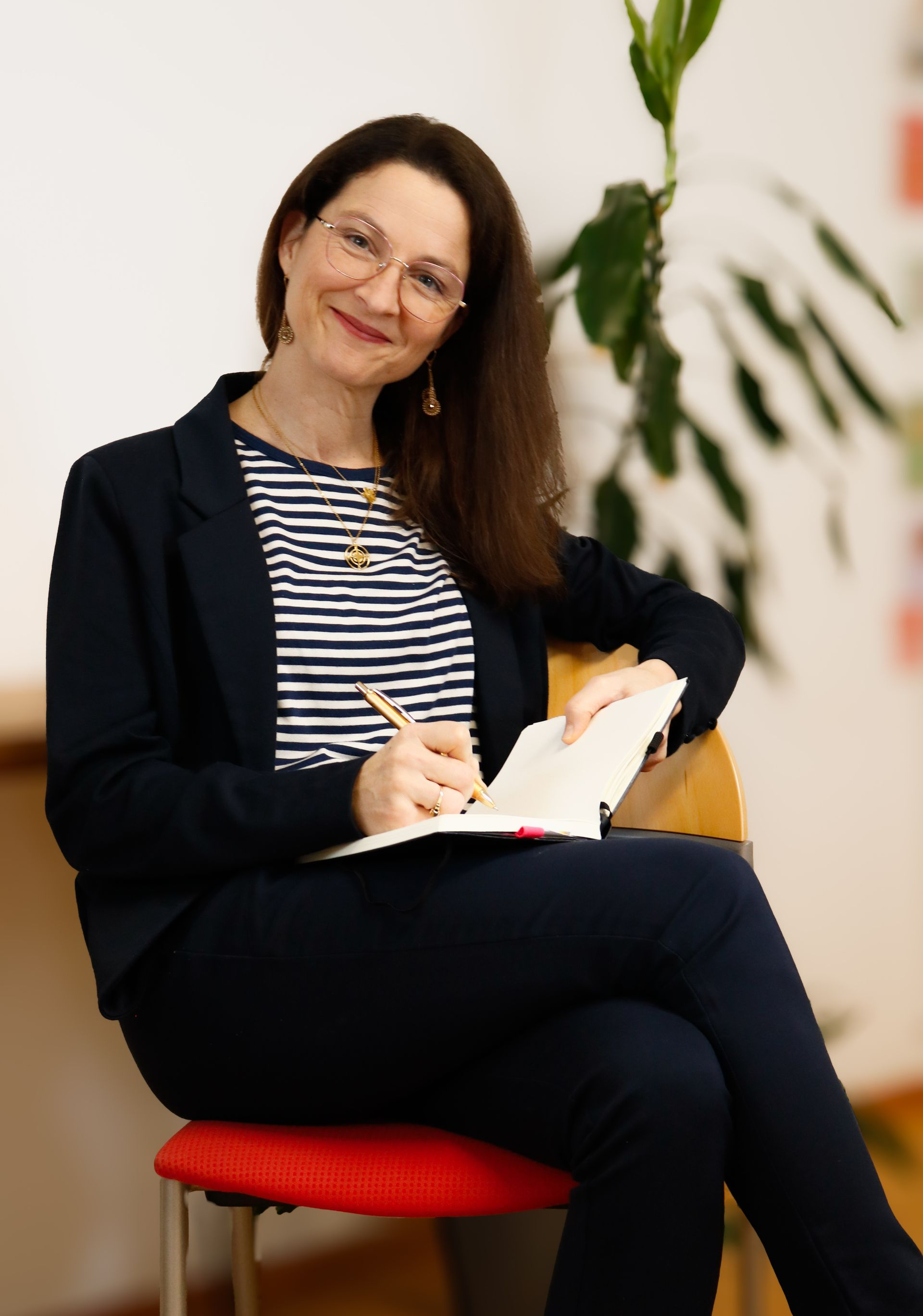 Eine Frau sitzt auf einem roten Stuhl, schreibt in ein Notizbuch und lächelt in einem hellen Innenraum.