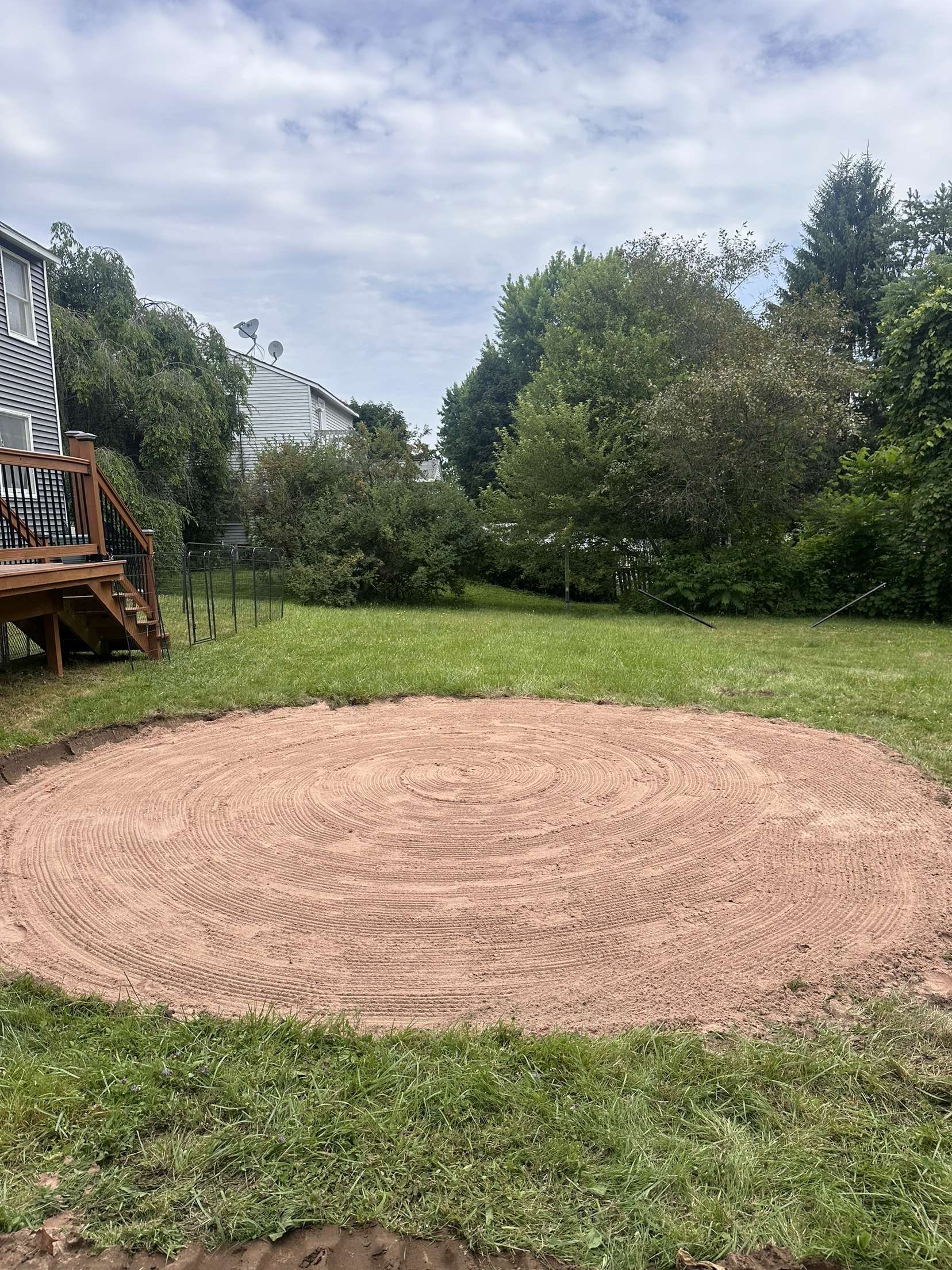 Oval sand pit on a green lawn, surrounded by grass and trees under a cloudy sky.