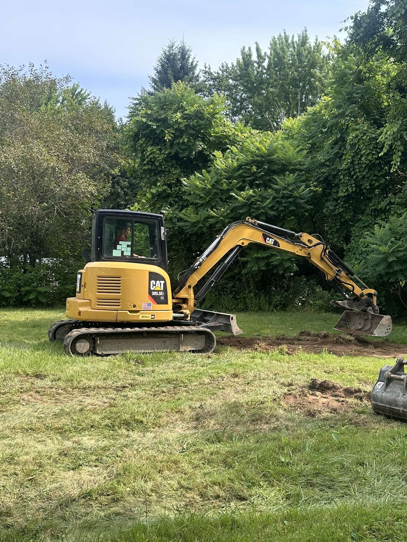 Yellow excavator digging in a grassy field, trees in background.