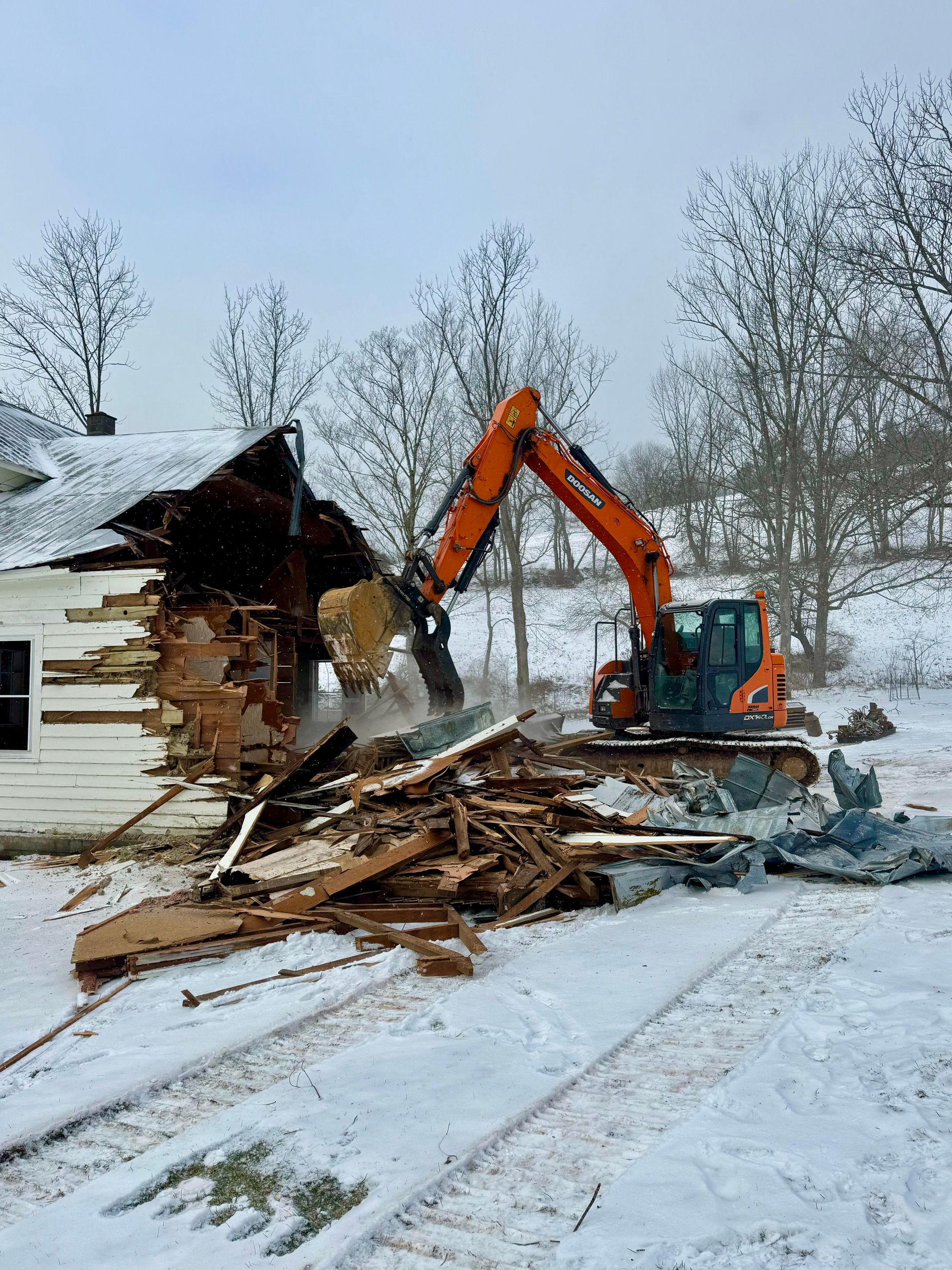 Excavator demolishing a house in a snowy, outdoor setting; orange machine, white house.