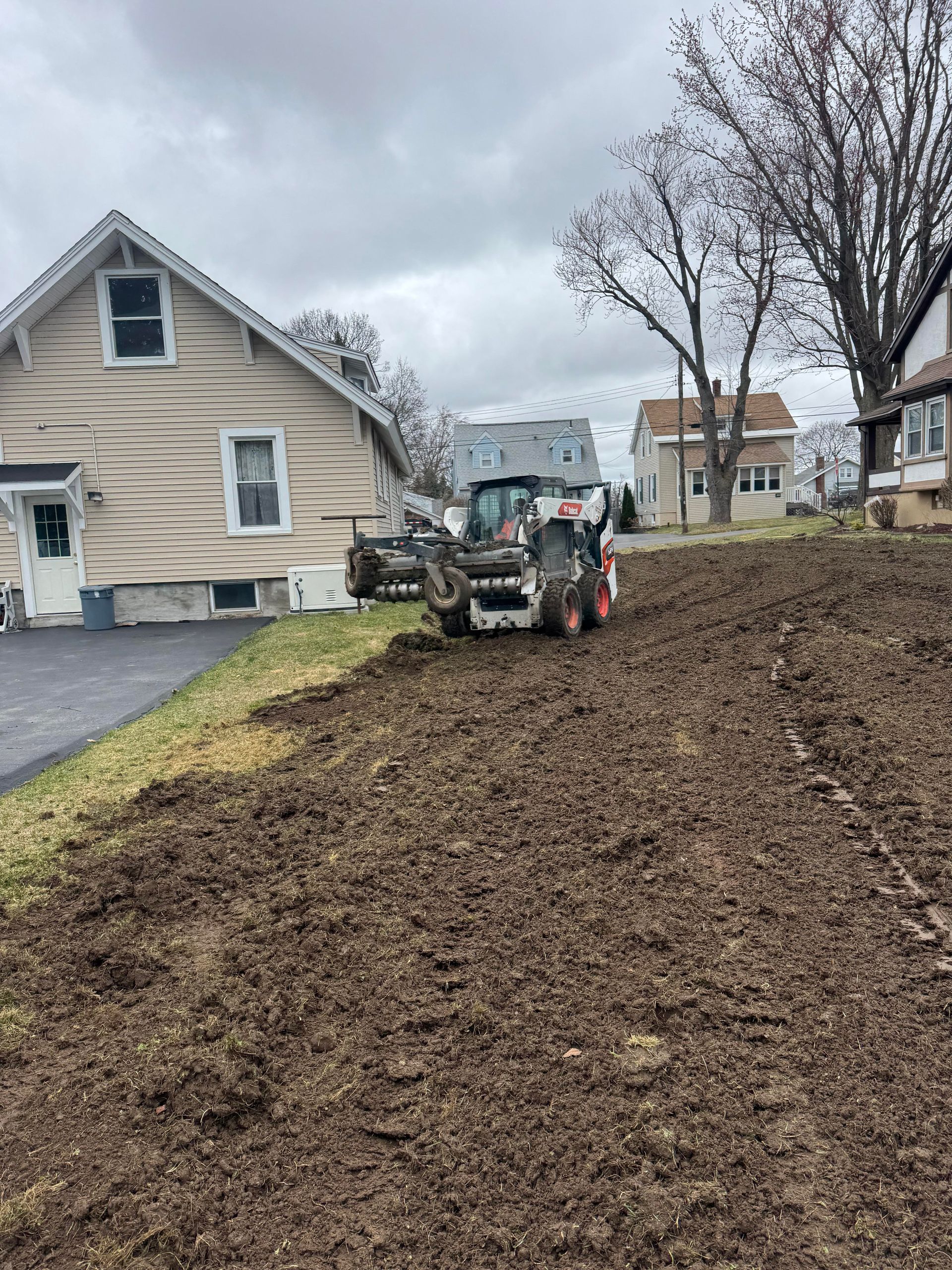 Skid steer tractor leveling muddy ground near houses under a cloudy sky.