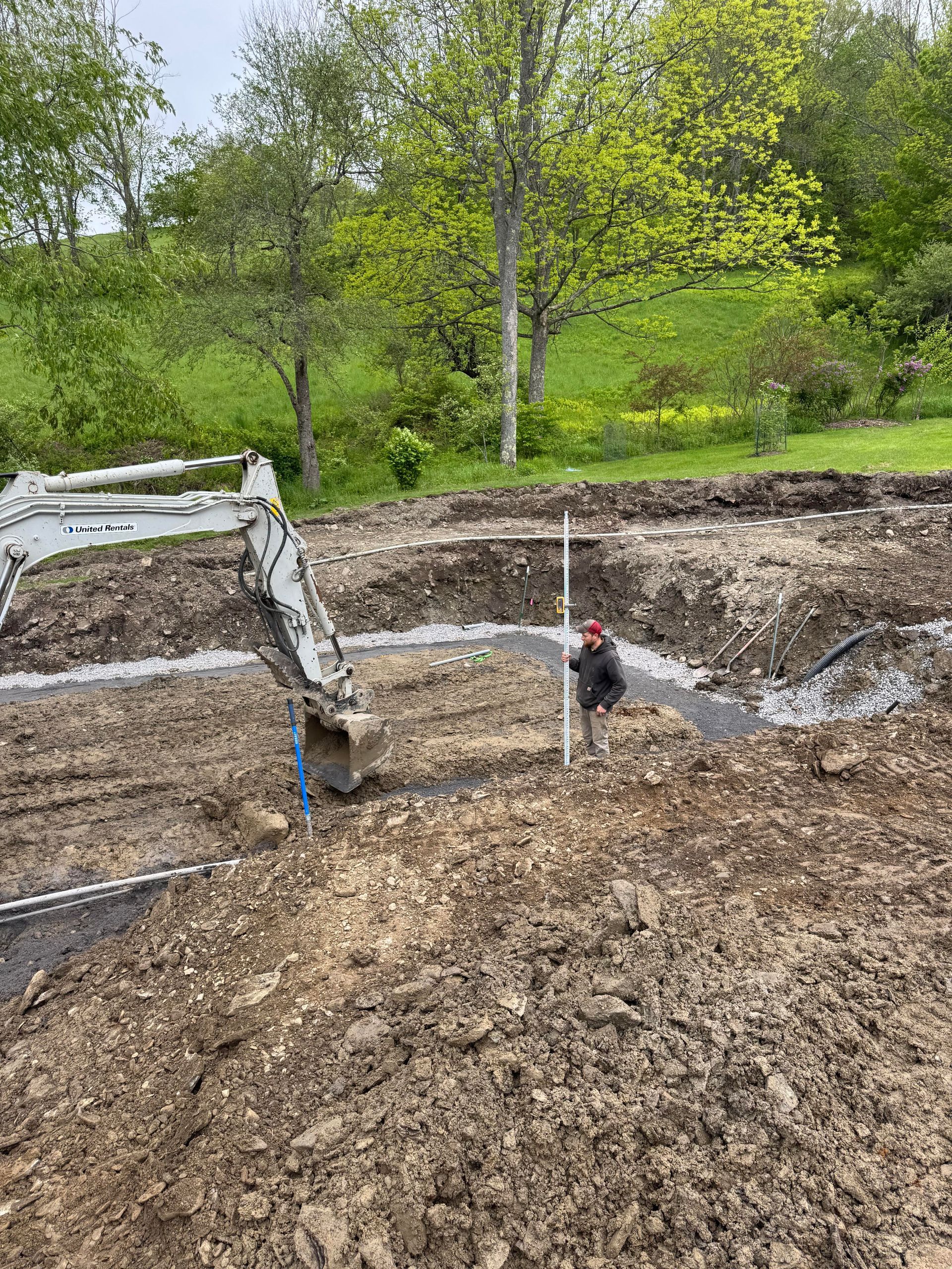 An excavator and worker in a dug-out area, preparing a construction site. Green trees and grass are in the background.