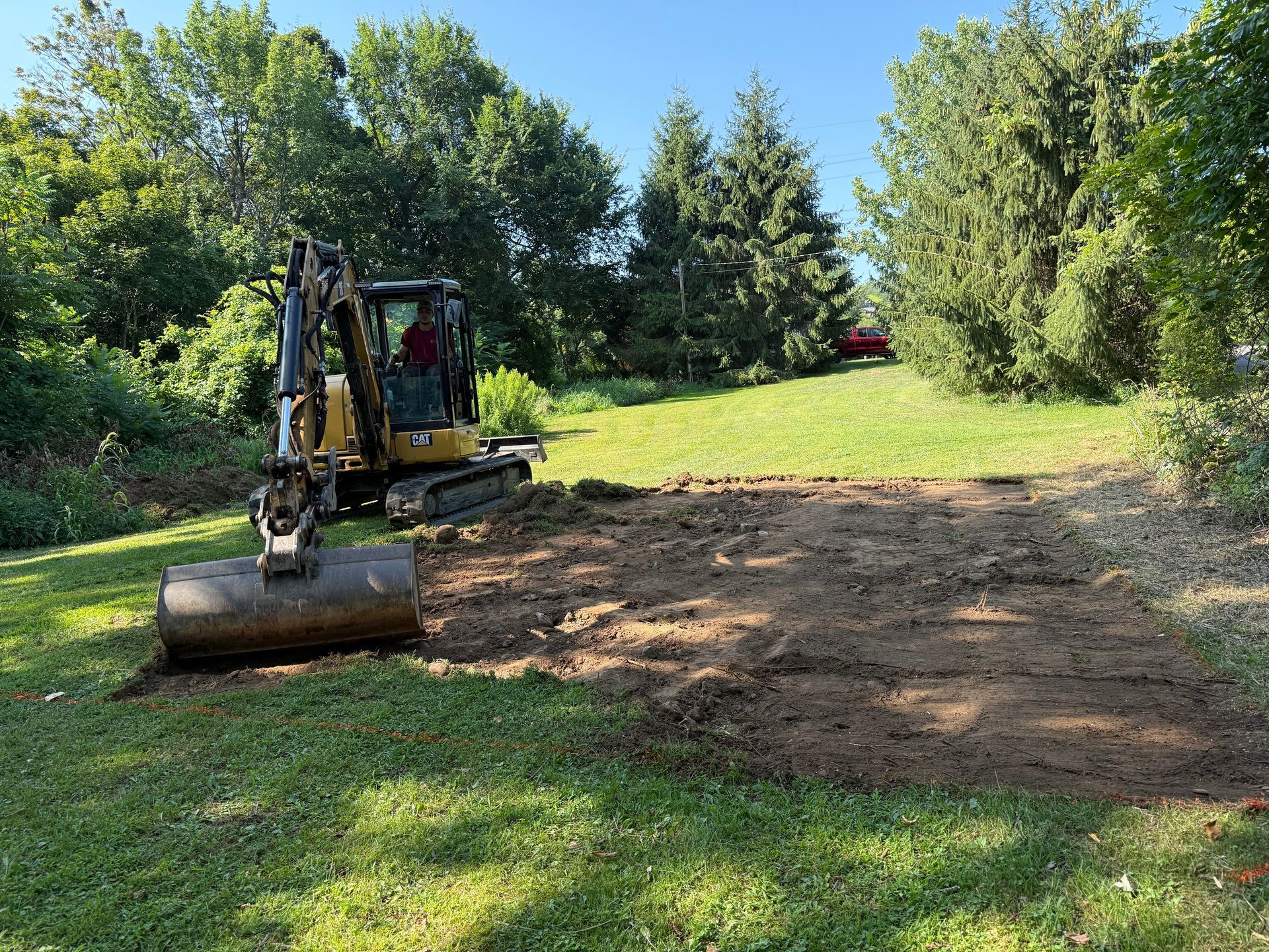 Mini excavator clearing a patch of dirt in a grassy area, trees in the background, blue sky.