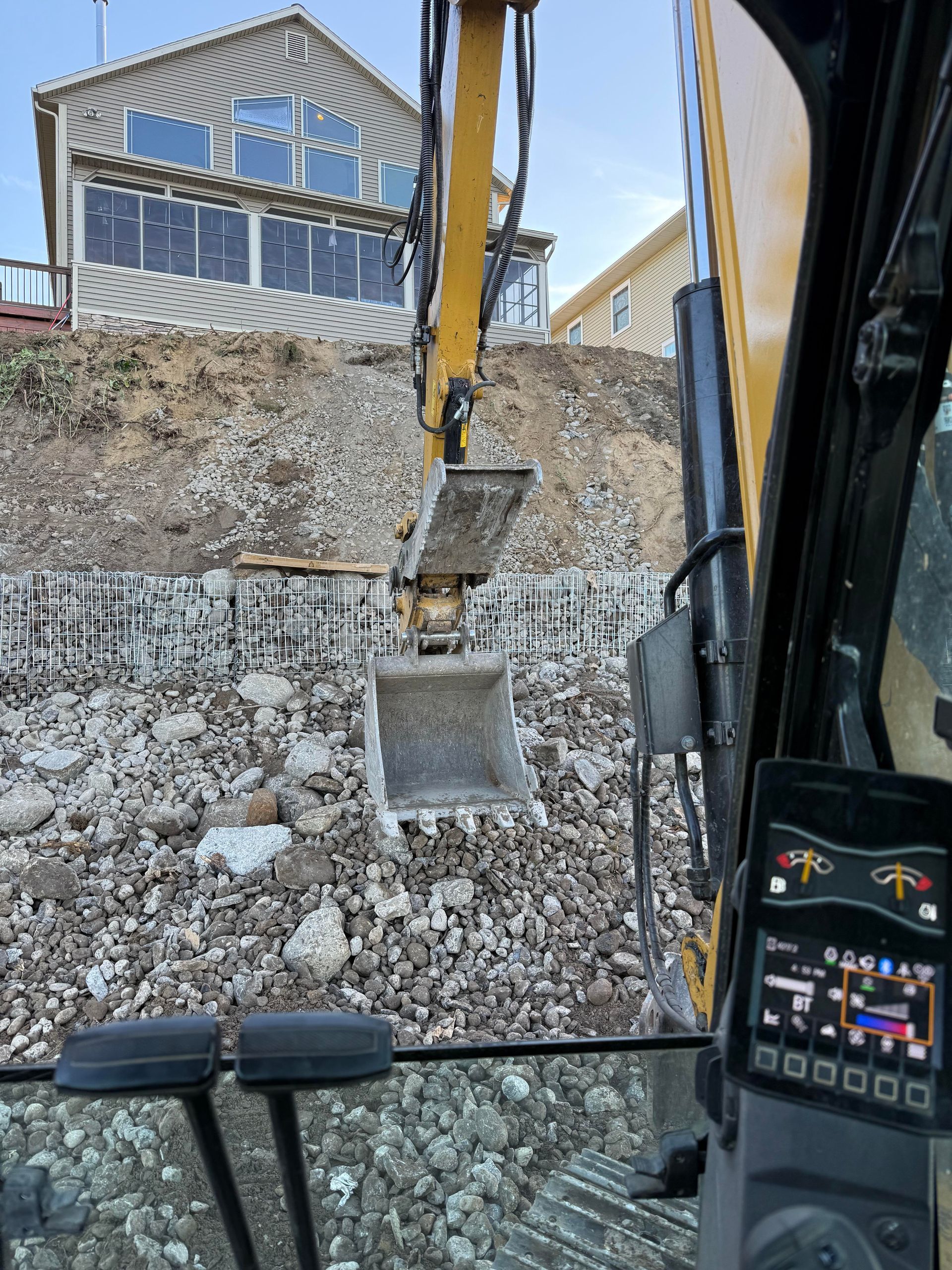 Excavator bucket scooping earth; building in background. View from inside the cab.
