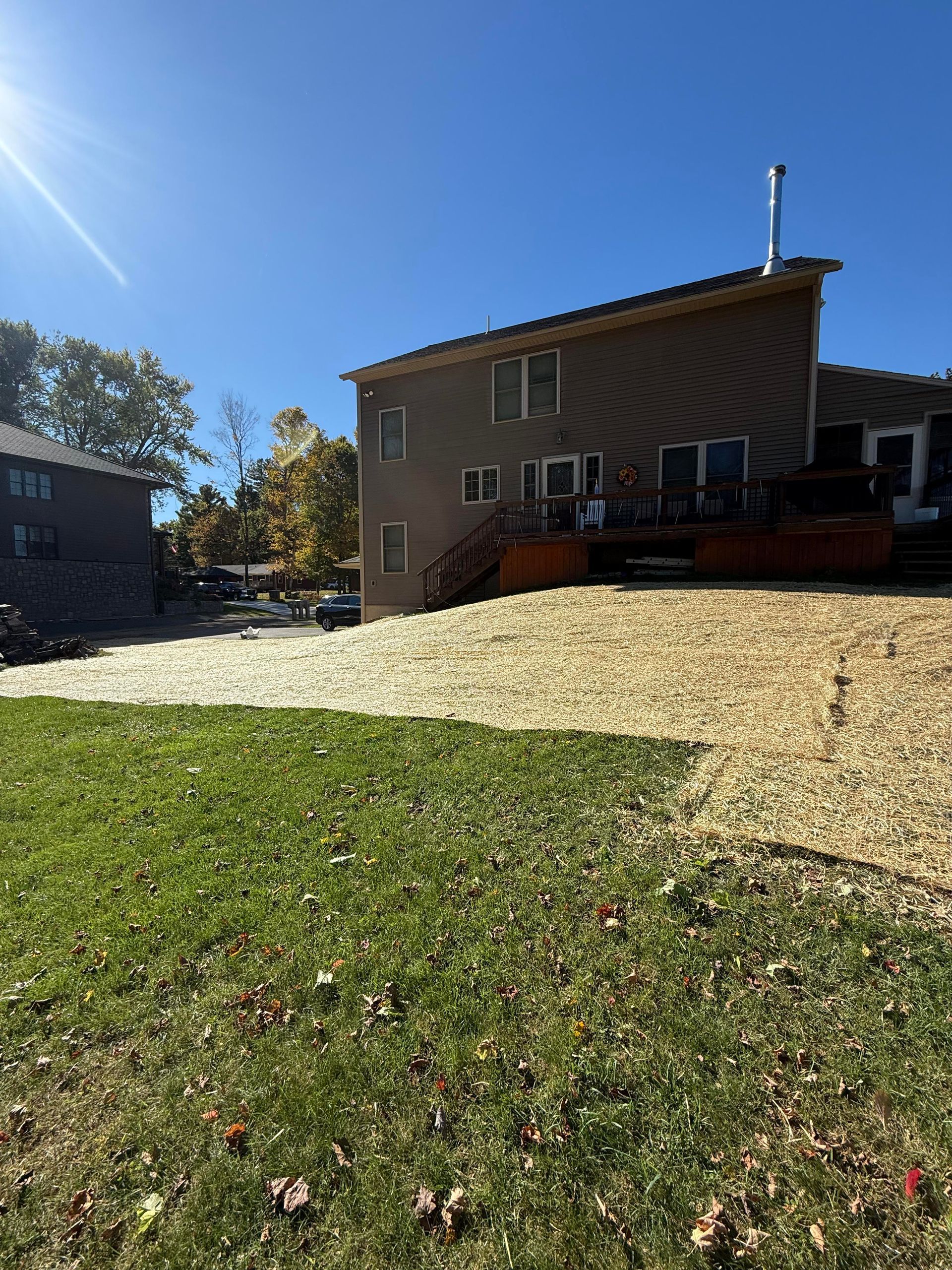 Backyard with green grass, wood chip ground cover, and a house with a deck on a sunny day.