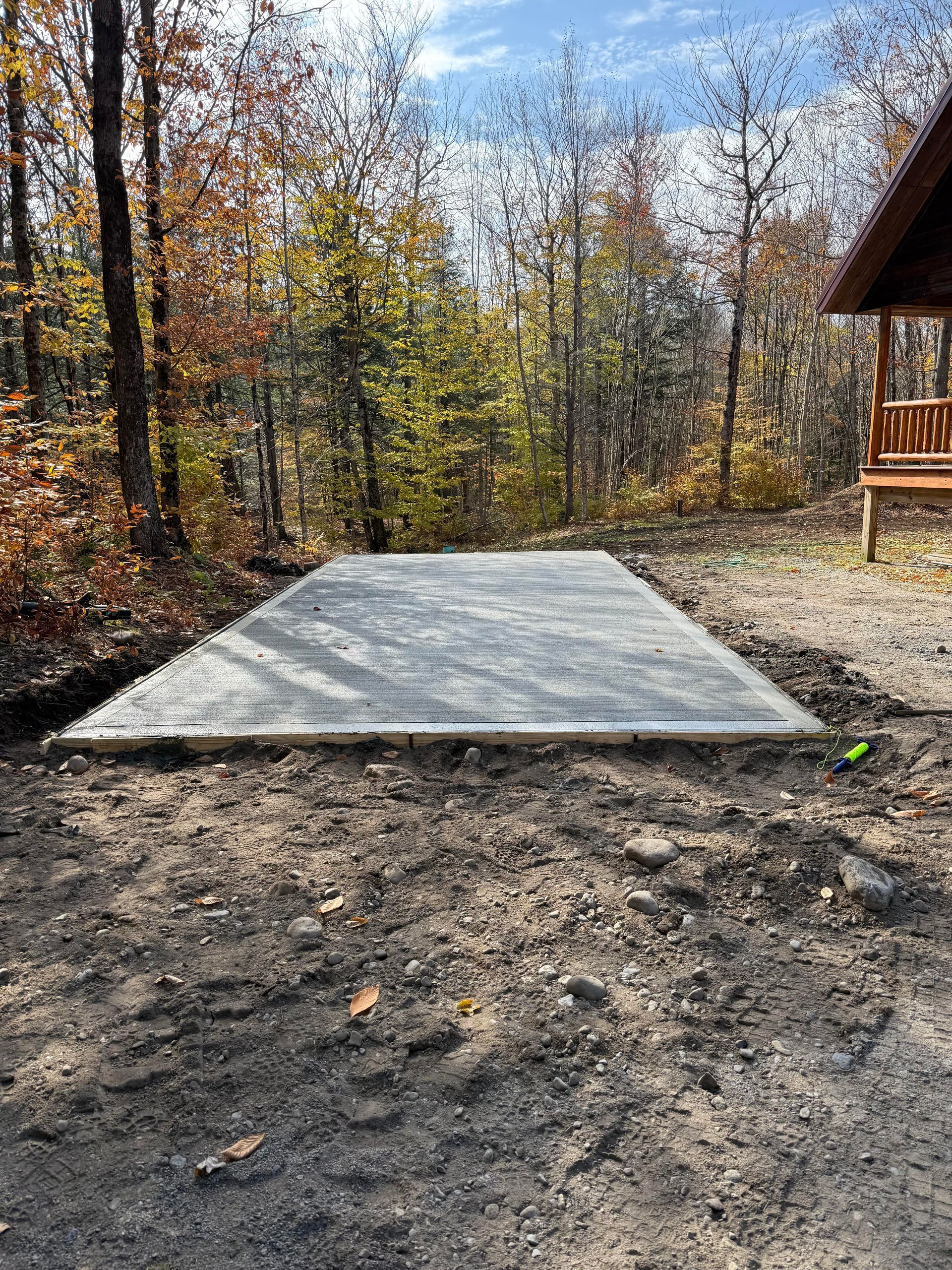 A newly poured concrete slab sits on dirt in a wooded area; a cabin with a porch is visible.
