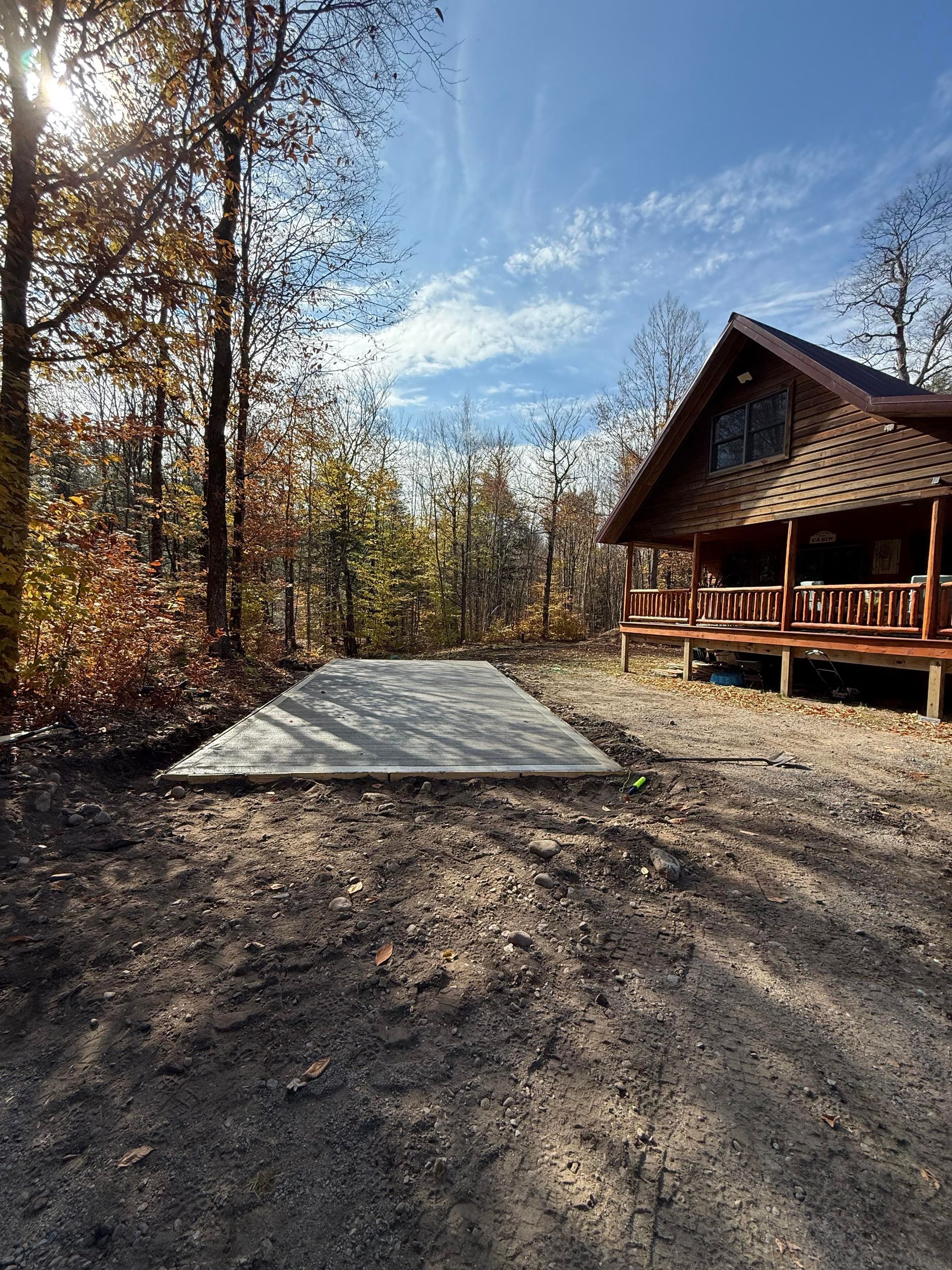 Concrete pad in a wooded area next to a log cabin, sunny day, blue sky.