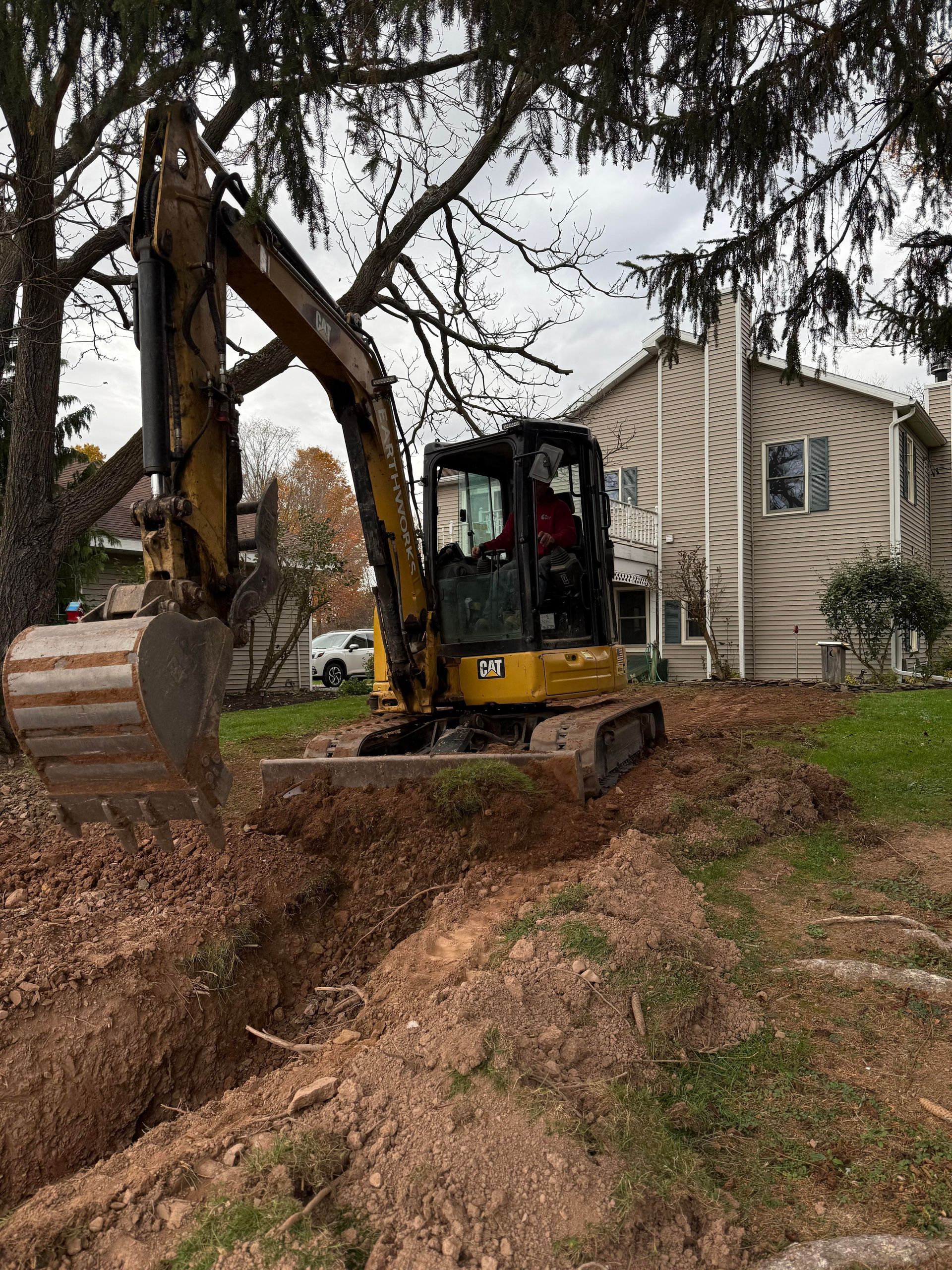 Yellow excavator digging a trench near a house with a person in the cab. Overcast sky.