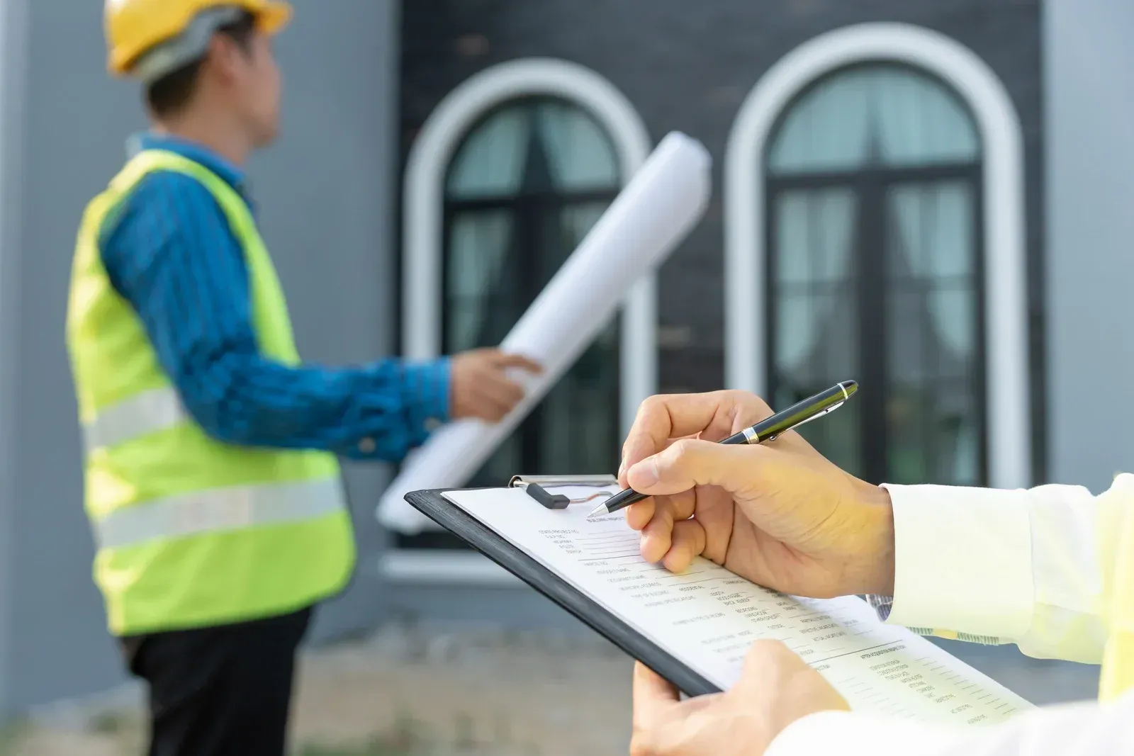 Construction worker writing on a clipboard, another holding blueprints outside a building with arched windows.