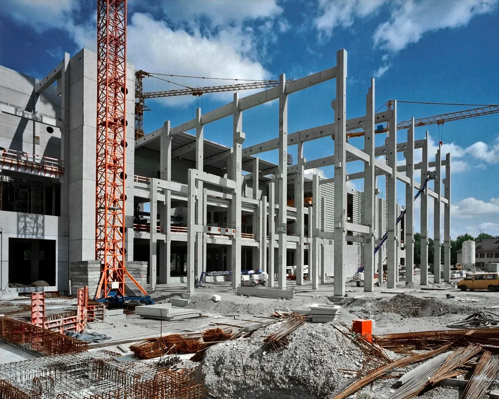 Construction site of a building with concrete frame, cranes, and building materials under a cloudy sky.