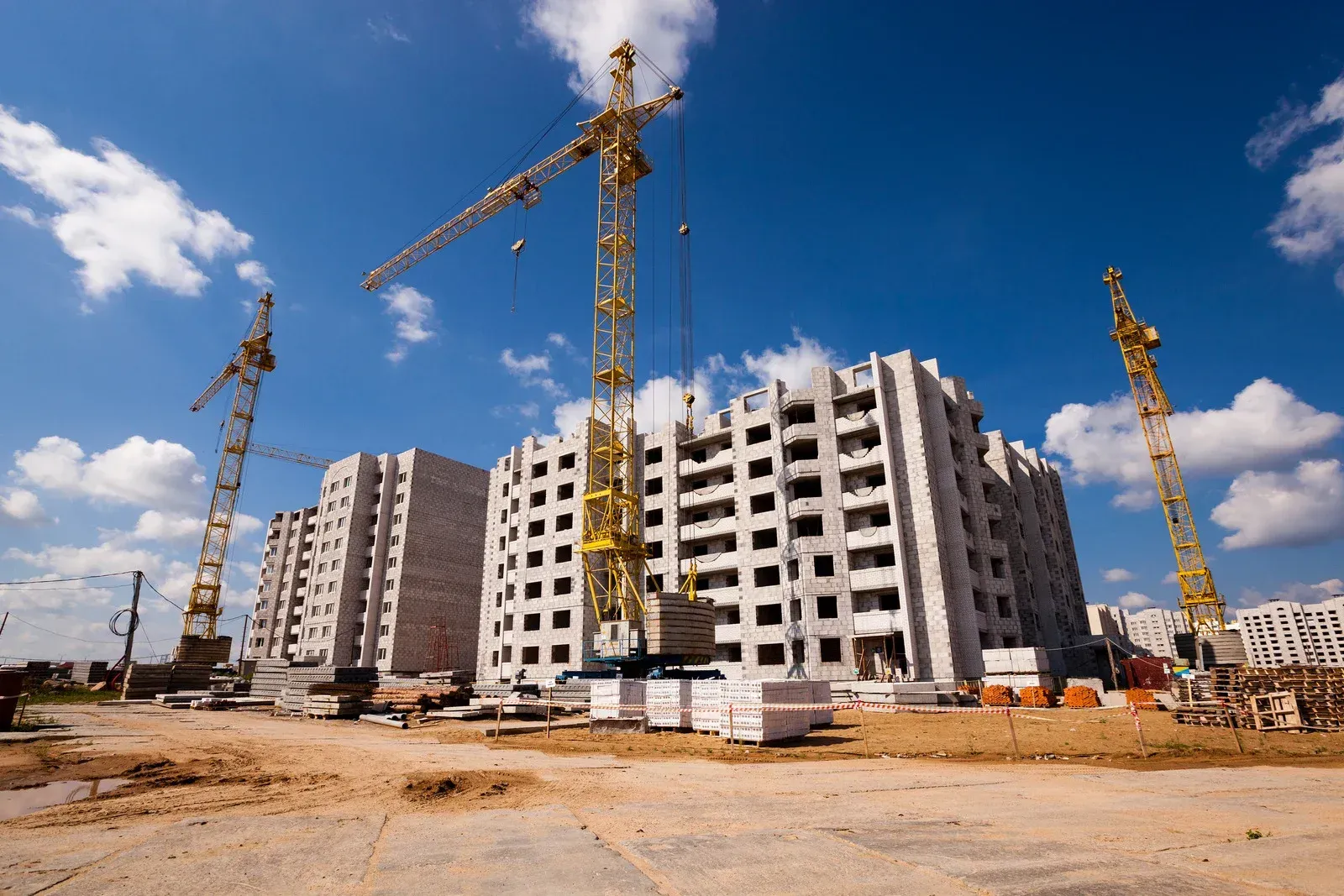 Construction site with tall yellow cranes, unfinished white concrete buildings, and blue sky.