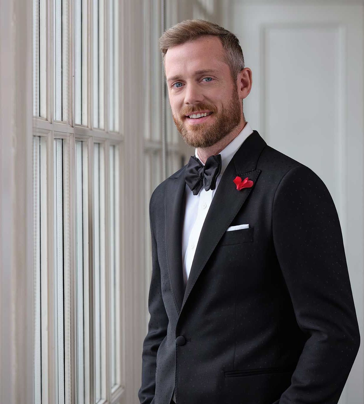 A man in a tuxedo and bow tie is standing in front of a window.