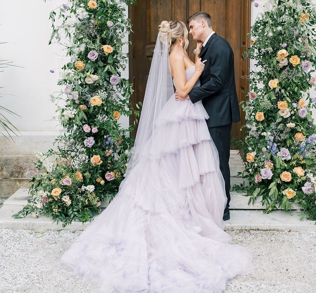 A bride and groom are kissing in front of a floral arch . the bride is wearing a purple dress.
