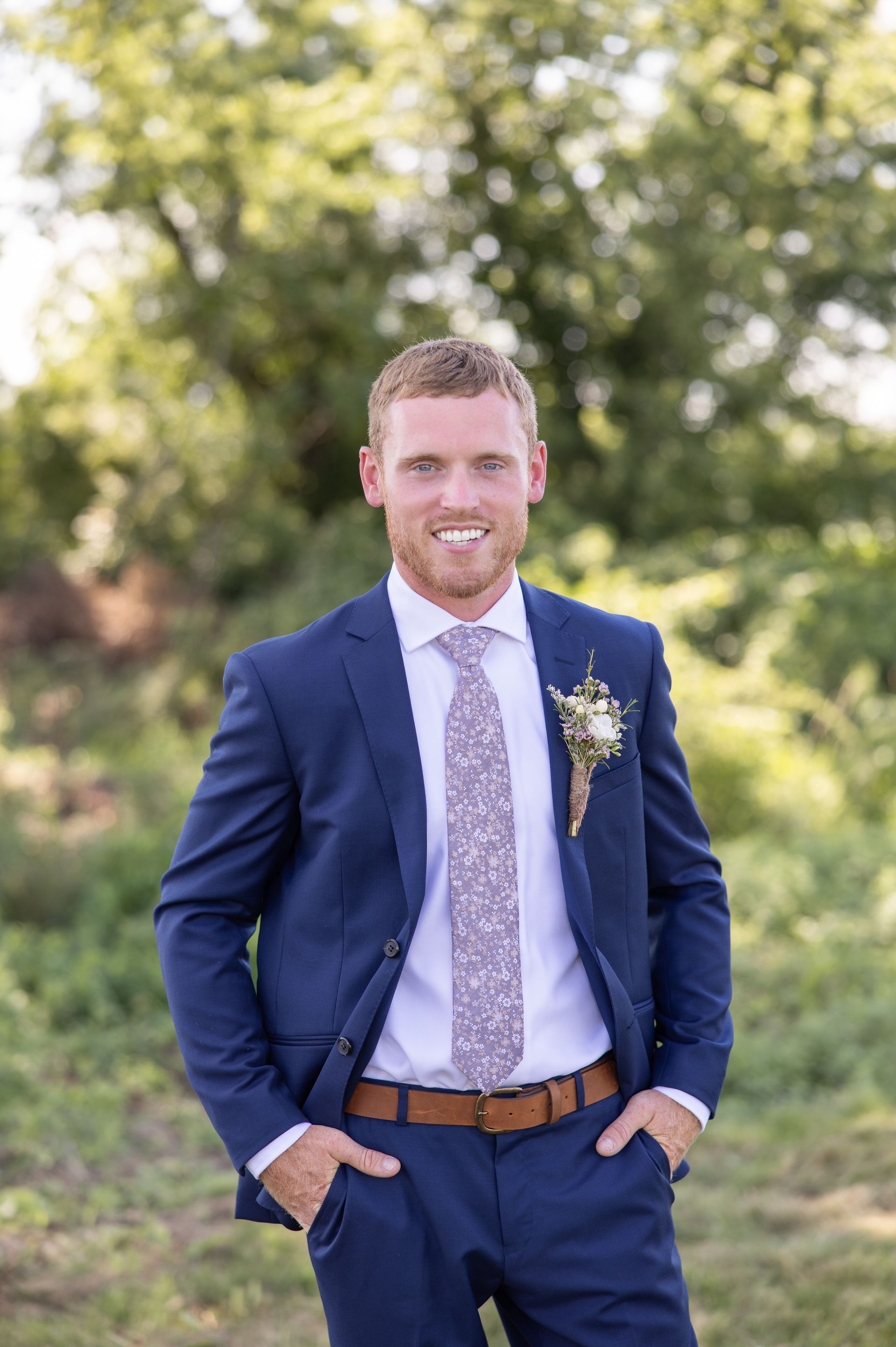 A man in a blue suit and tie is standing in a field with his hands in his pockets.