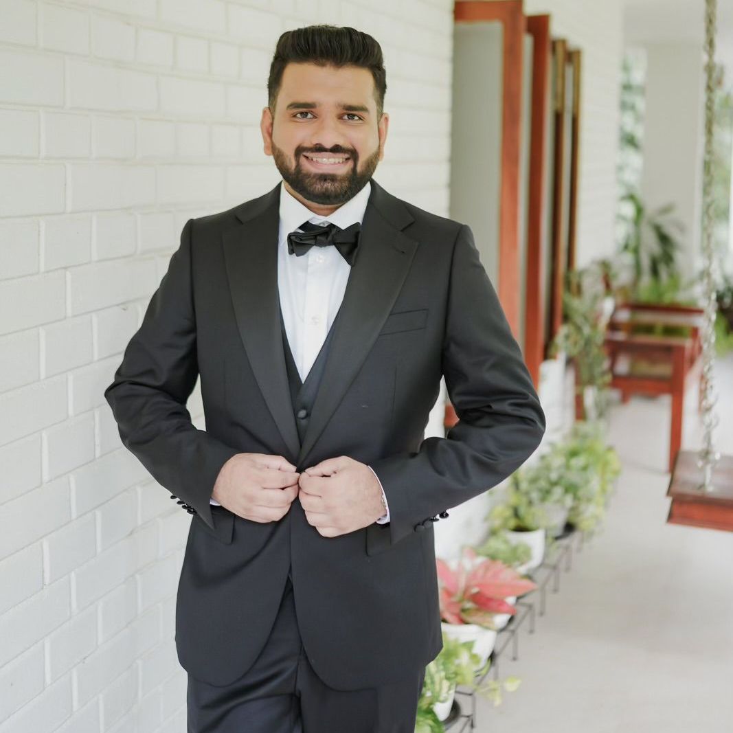 A man in a tuxedo and bow tie is standing in front of a white brick wall.