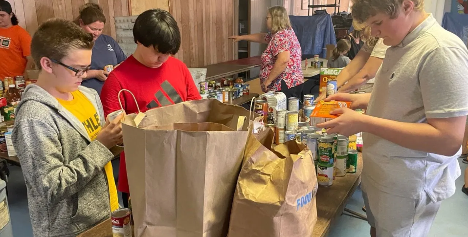 A group of young Boy Scouts are standing around a table sorting bags of food.