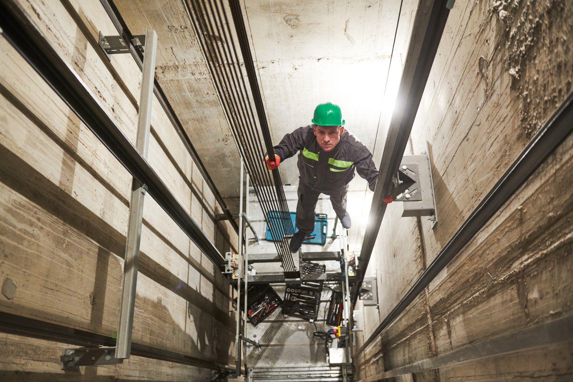 Elevator repairman in green helmet and work clothes, working inside an elevator shaft.