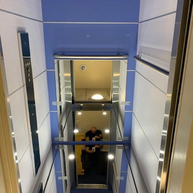 Elevator shaft interior, looking down. A person is sitting in the elevator car. Blue and white walls, metal accents.