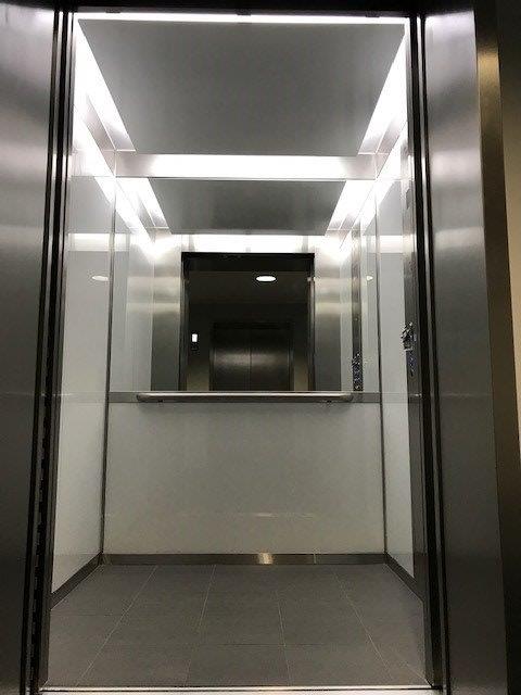 Interior view of a modern elevator with white walls, stainless steel accents, and overhead lighting.