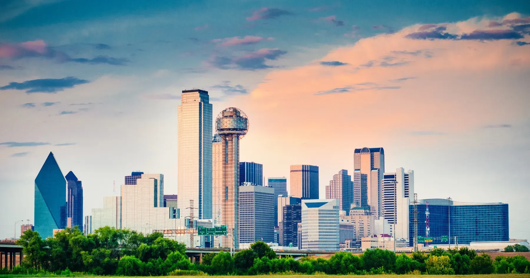 City skyline at sunset with modern skyscrapers, a skyline tower, and green trees in the foreground.
