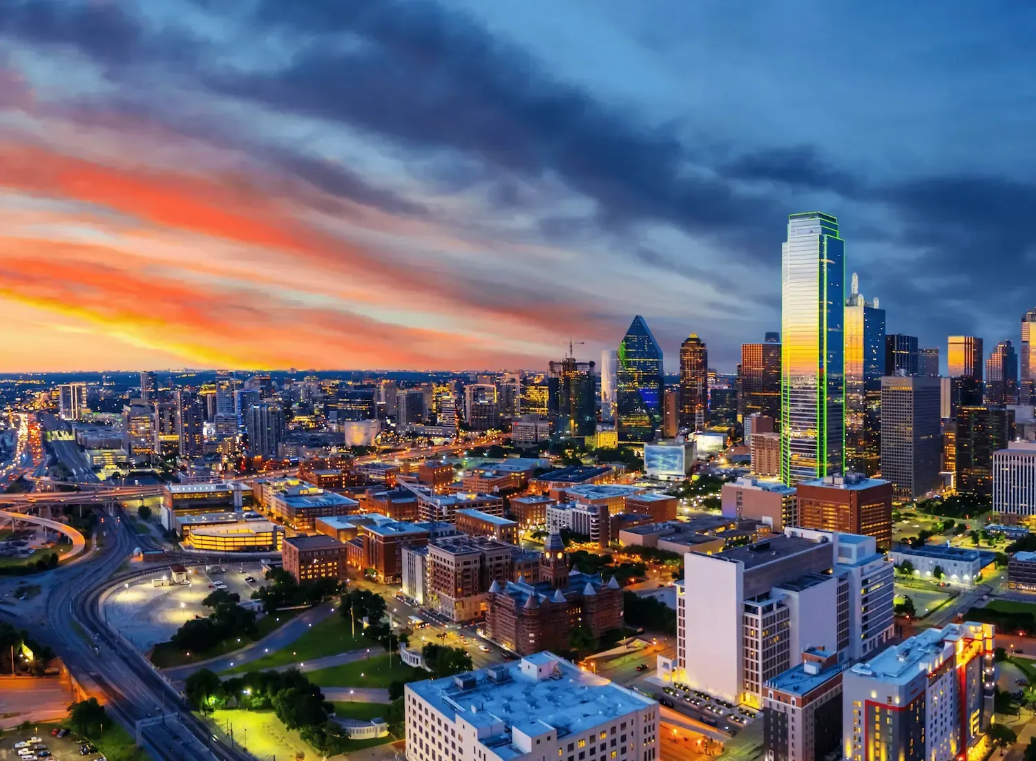 City skyline at sunset with colorful clouds and illuminated skyscrapers overlooking a river.