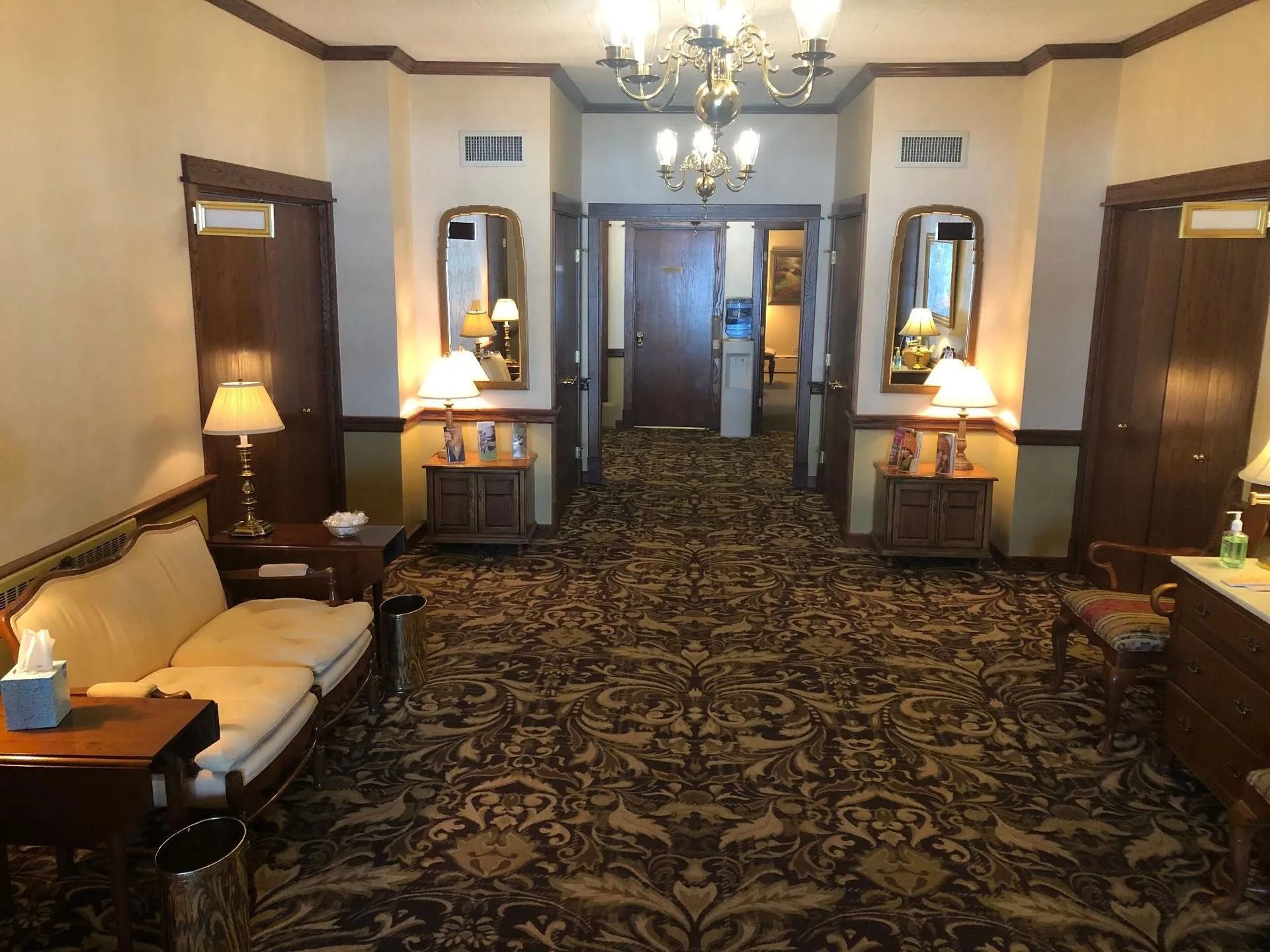 An elegant hotel lobby or hallway with patterned carpets, two upholstered benches, wall mirrors, lamps, and wood doors.