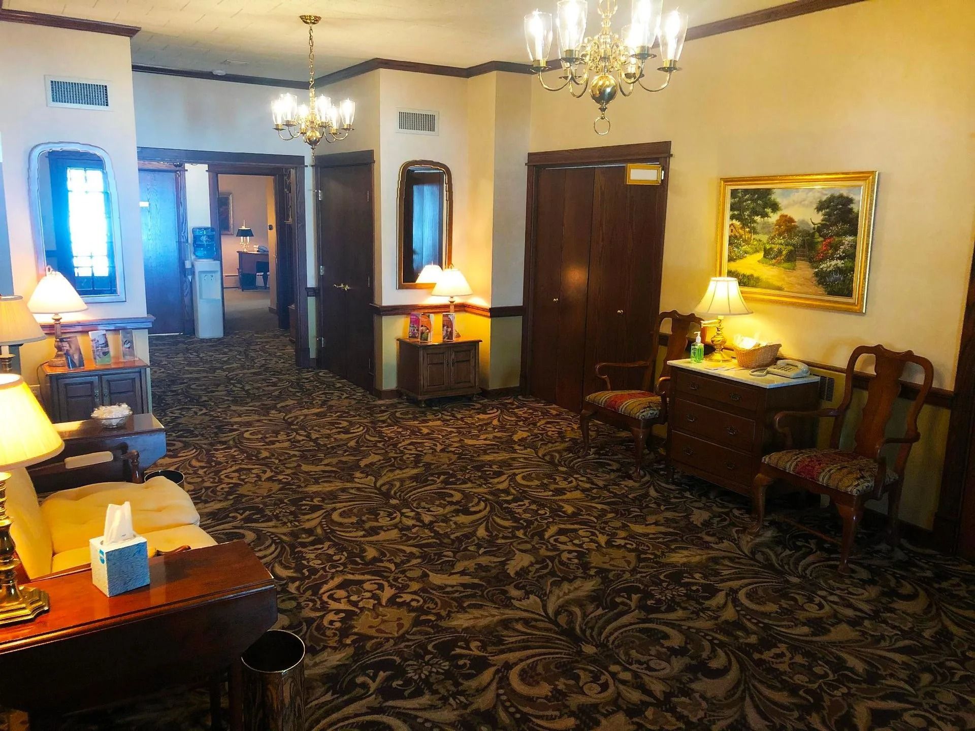 A dimly lit, carpeted hotel lobby or hallway with wood doors, antique furniture, chandeliers, and framed artwork.