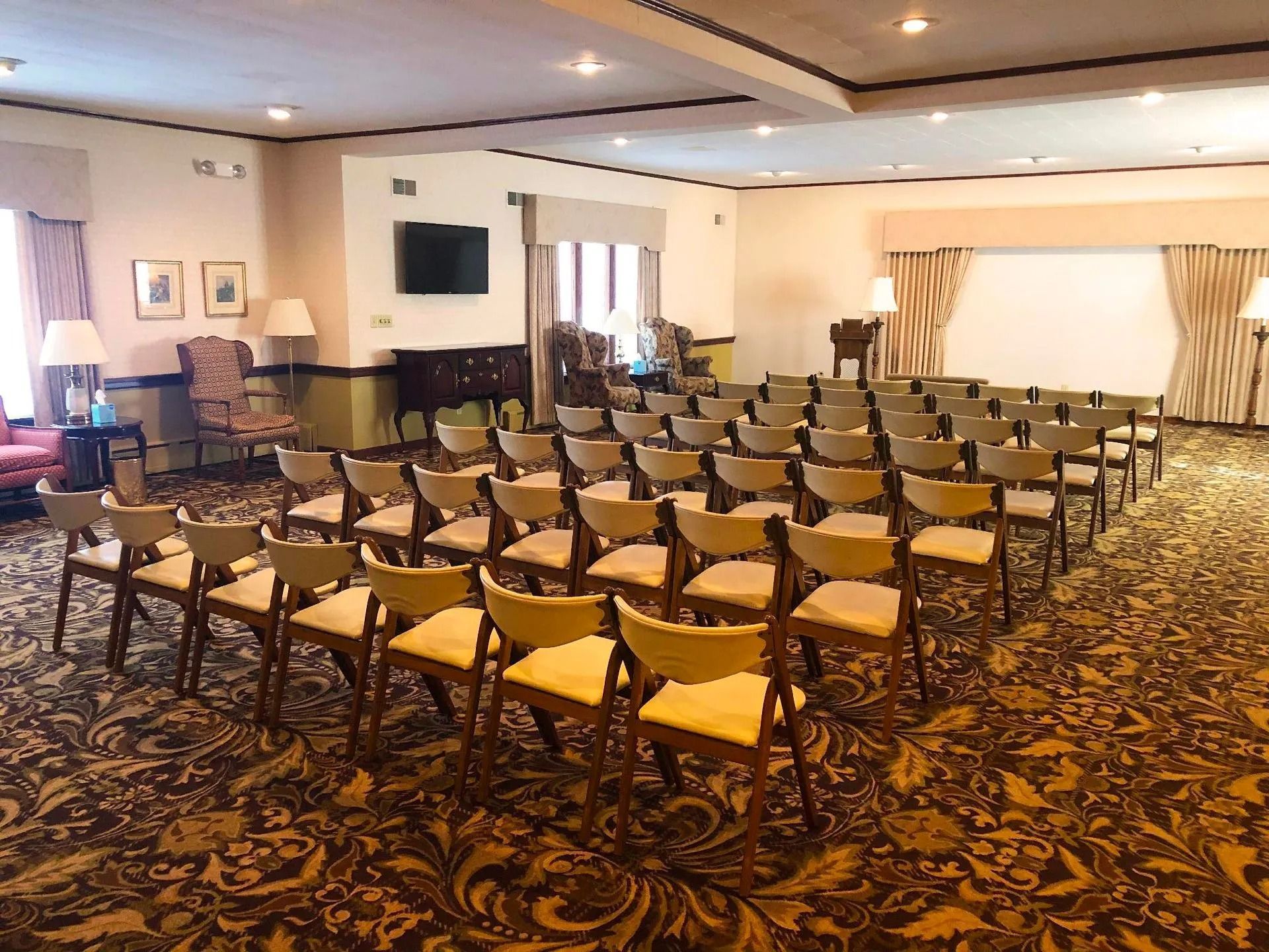 A conference room with rows of empty beige chairs facing an open area, set against a patterned gold and brown carpet.