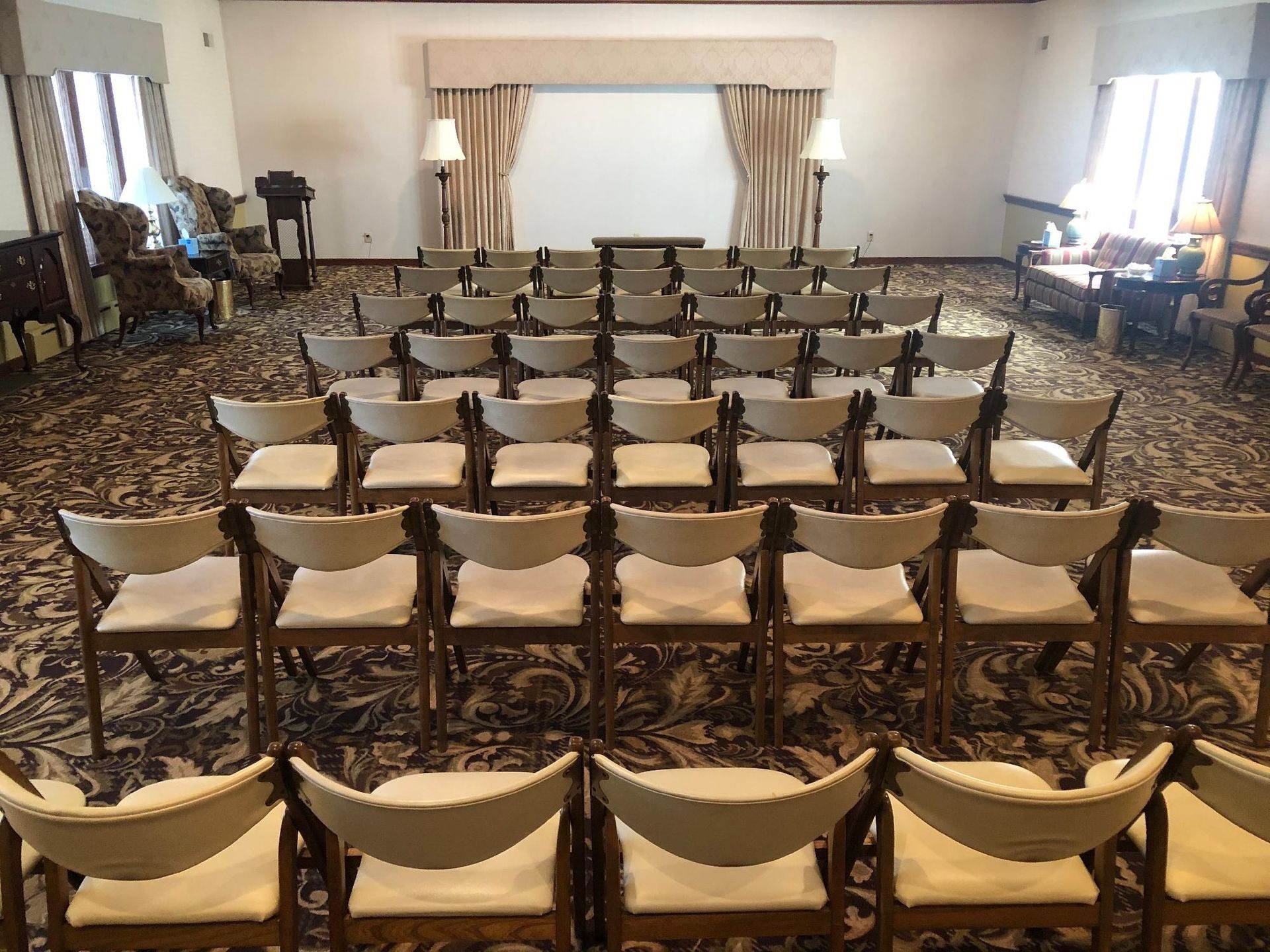Rows of chairs arranged for an event in a carpeted room with curtains and lamps at the front.