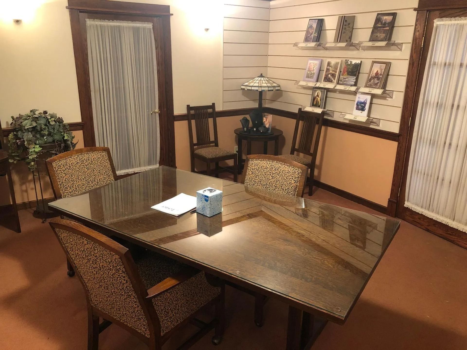 A formal meeting room with a large glass table, four patterned chairs, a lamp, and wall-mounted brochures.