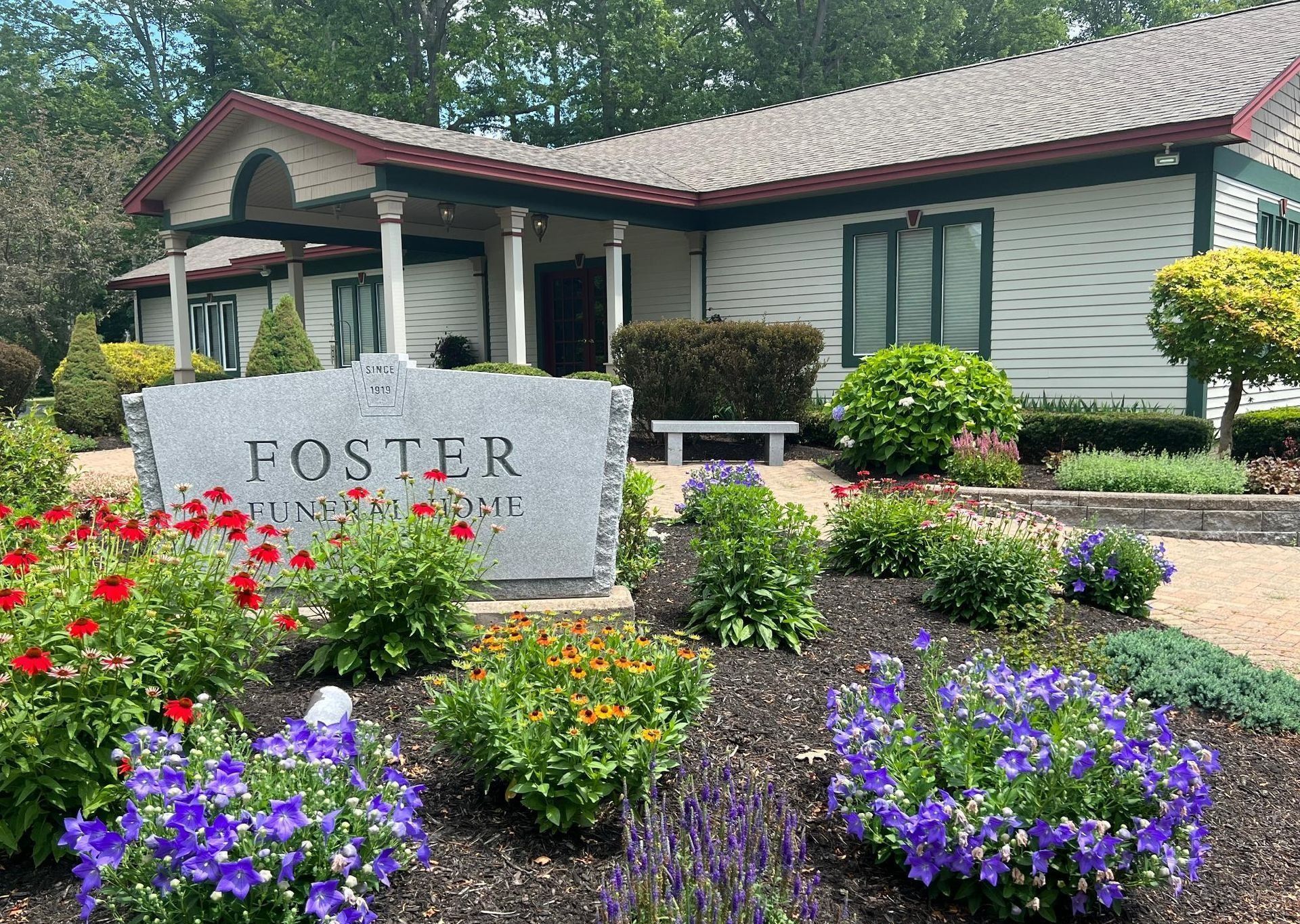 A stone sign for Foster Funeral Home sits in front of a white building surrounded by blooming flowers and green bushes.