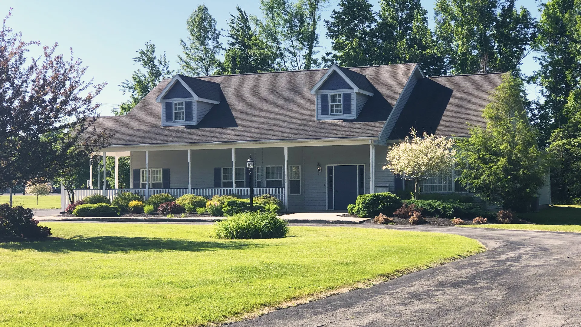 A grey, single-story house with two dormer windows, a long front porch, and a gravel driveway in a grassy, rural setting.