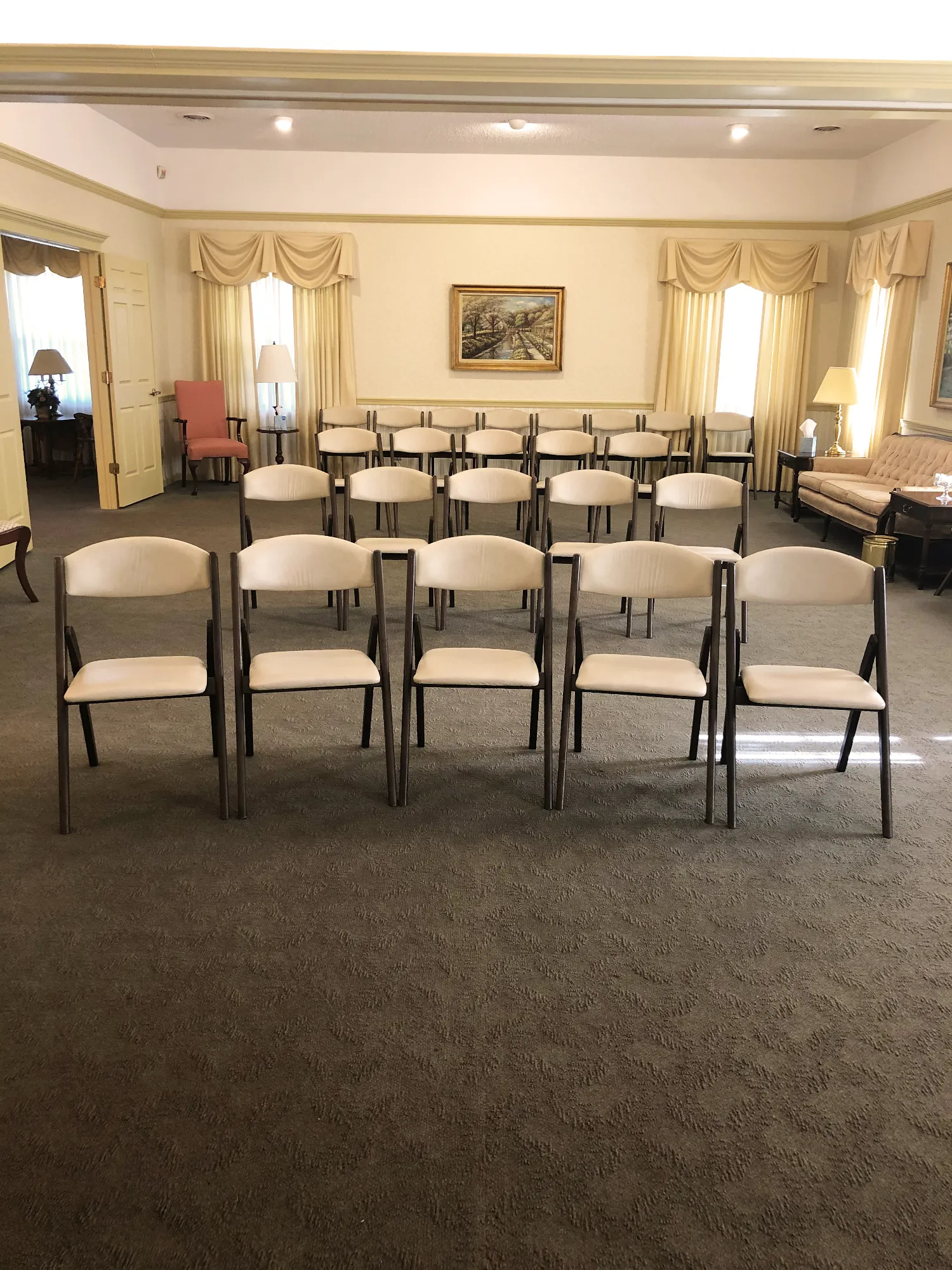 A room set up with rows of folding chairs facing a wall with a framed picture, flanked by windows with curtains.