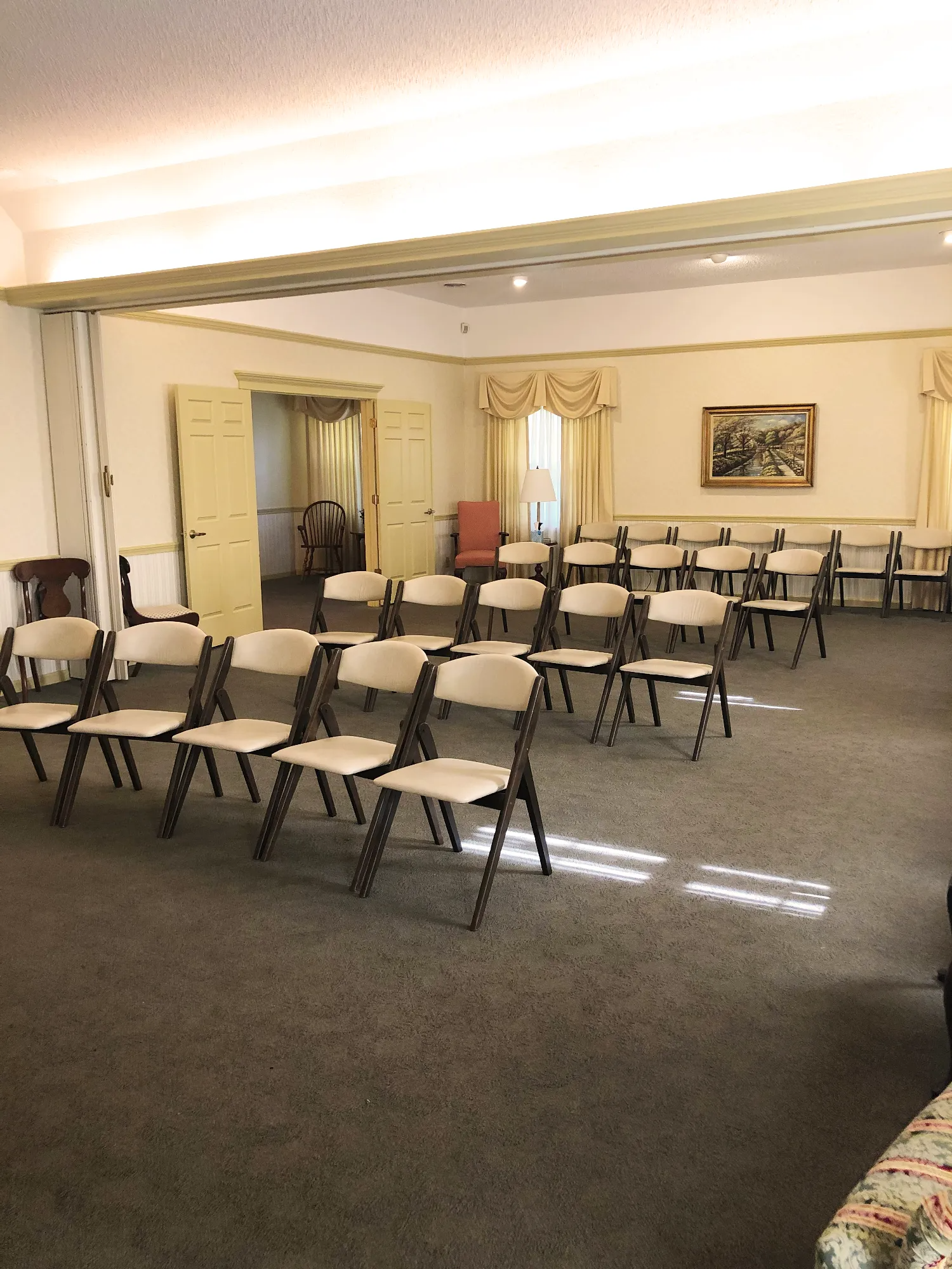 A room with rows of light-colored folding chairs arranged on a carpeted floor facing a wall with a framed picture.