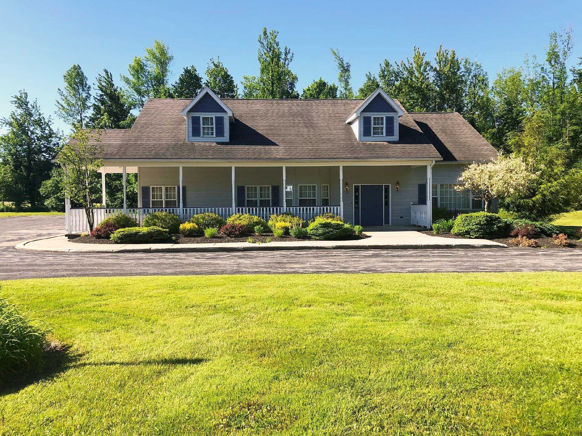 A two-story tan house with a wrap-around porch, blue shutters, and two dormers, situated behind a grassy lawn and gravel.
