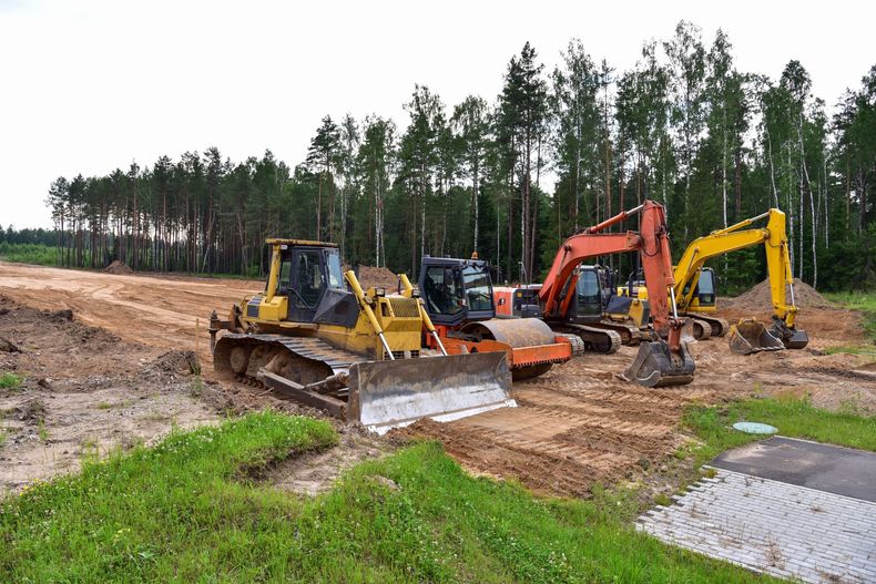 Construction site with bulldozers and excavators on a dirt lot beside a wooded area