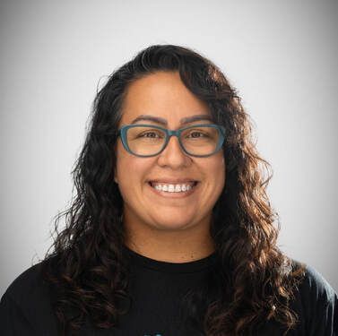 Smiling person with curly dark hair and glasses against a light gray background