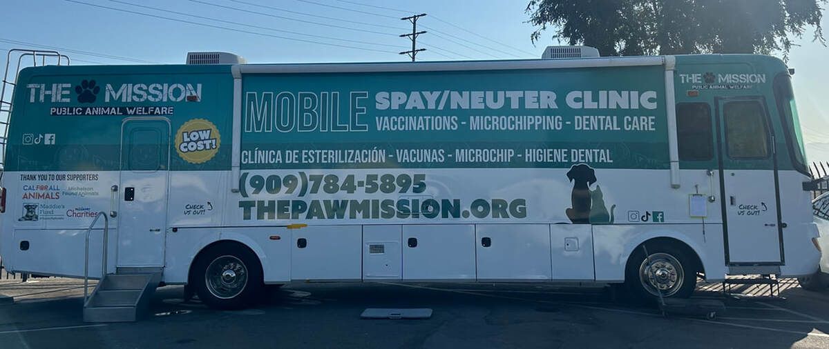 White mobile clinic van for The Mission parked outdoors with large teal signage and phone numbers.
