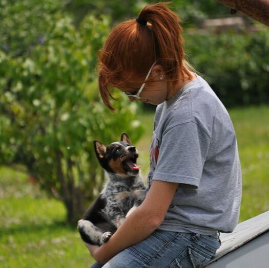 A woman in a gray T-shirt sits outdoors holding a small dog that looks up at her.