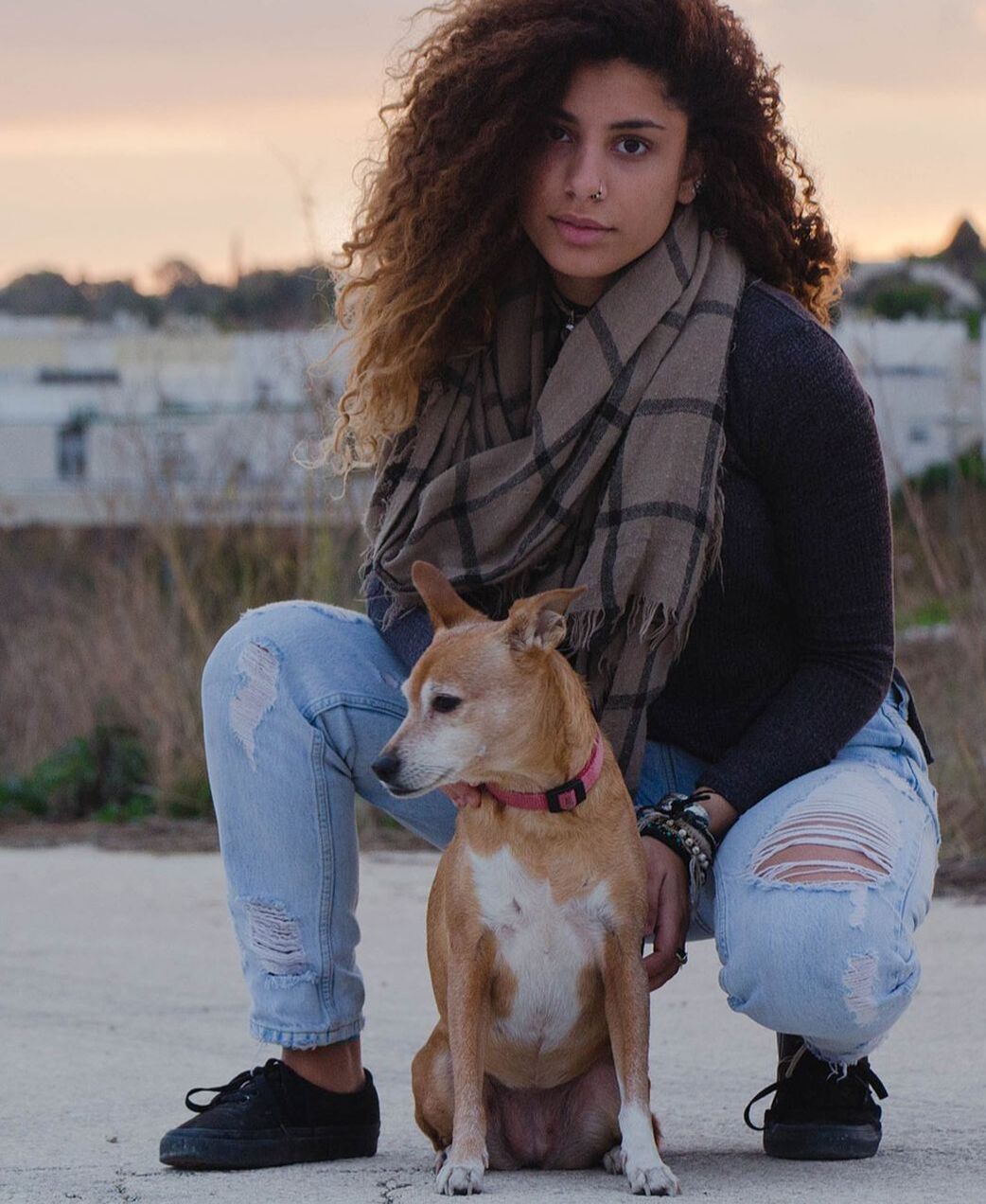 Woman crouches with a tan dog on a paved path at sunset, wearing ripped jeans and a plaid scarf.