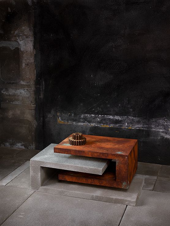 Een geometrische salontafel in roestbruine en grijze tinten met een tandwielachtig sculptuur tegen een donkere, gestructureerde muur.