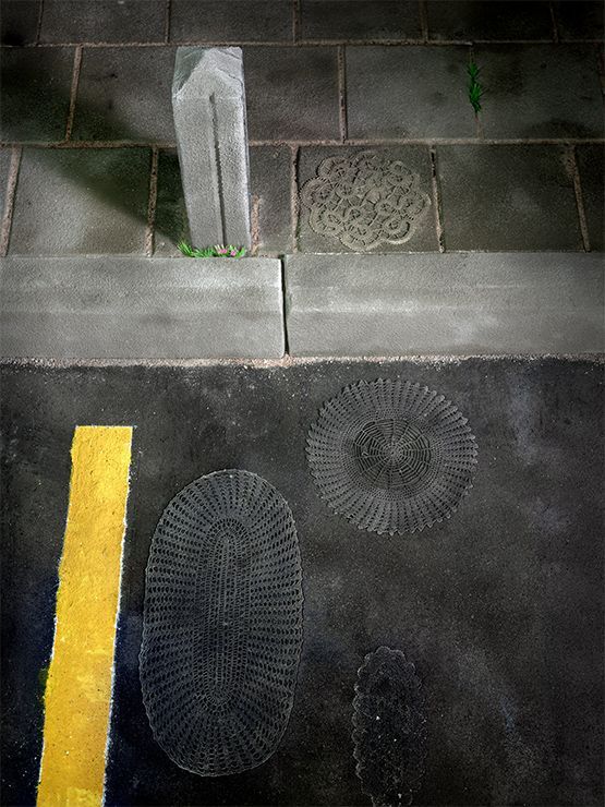 Stoep, straat en gele lijn met metalen putdeksels met structuur. Er staat ook een betonnen paal.
