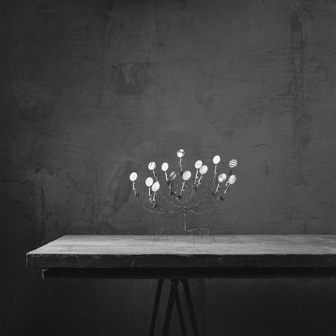 Metalen sculptuur van lepels met reflecterende lampjes op een tafel met textuur tegen een donkere muur.
