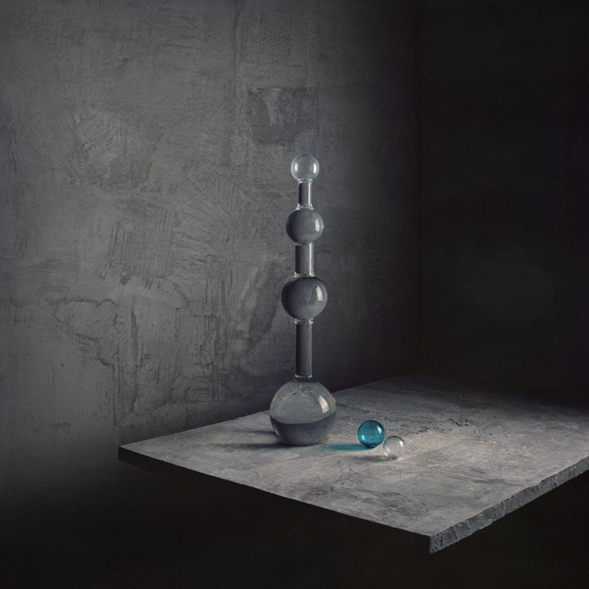 Een hoge, grijze glazen sculptuur en een blauwe bol staan ​​op een betonnen plank tegen een grijze muur met structuur.