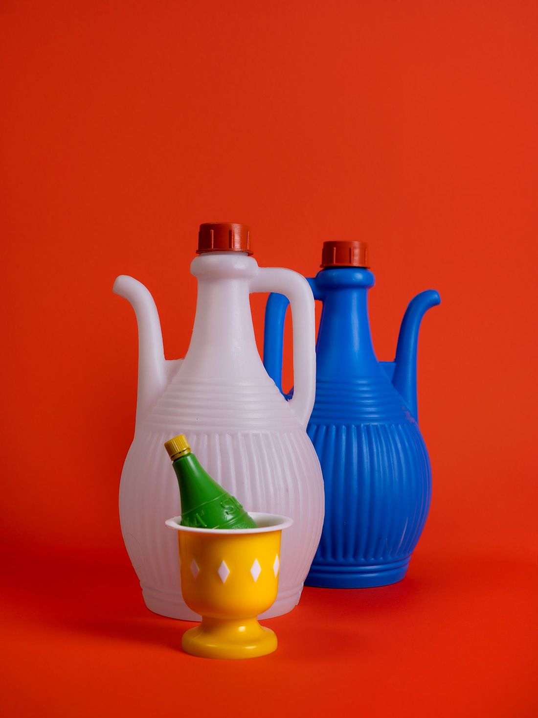 Twee plastic kannen (wit en blauw) en een gele beker met een groene fles op een rode achtergrond.