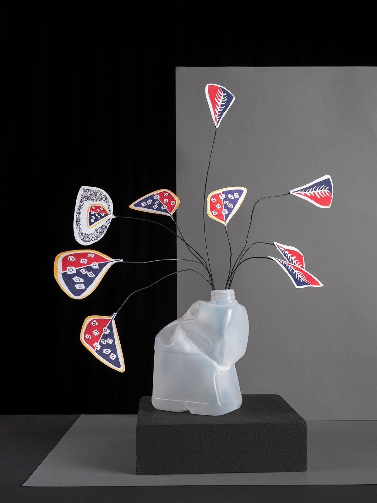 Sculptuur van papieren vlaggen met rode en blauwe patronen aan draden in een vaas van een verfrommelde plastic fles, met een zwarte achtergrond.