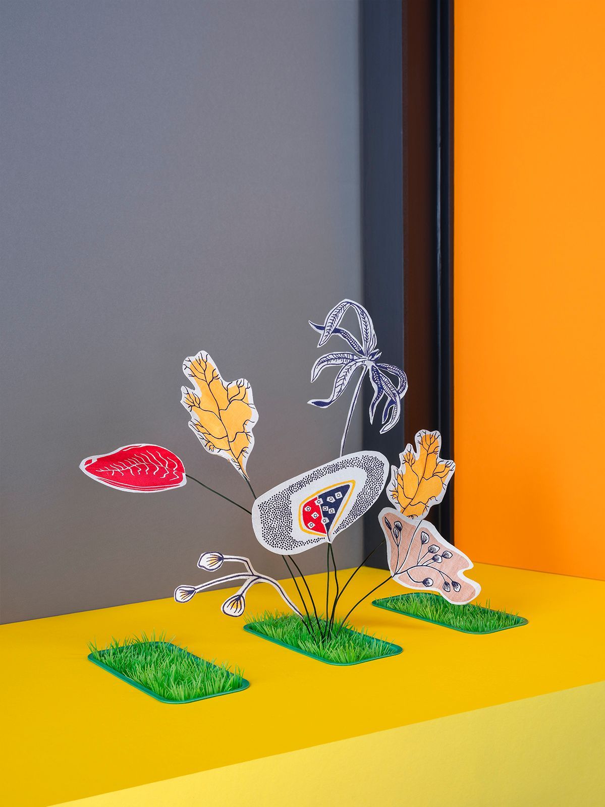 Kleurrijke papieren bloemen op kunstgras, tegen een grijze en oranje achtergrond.